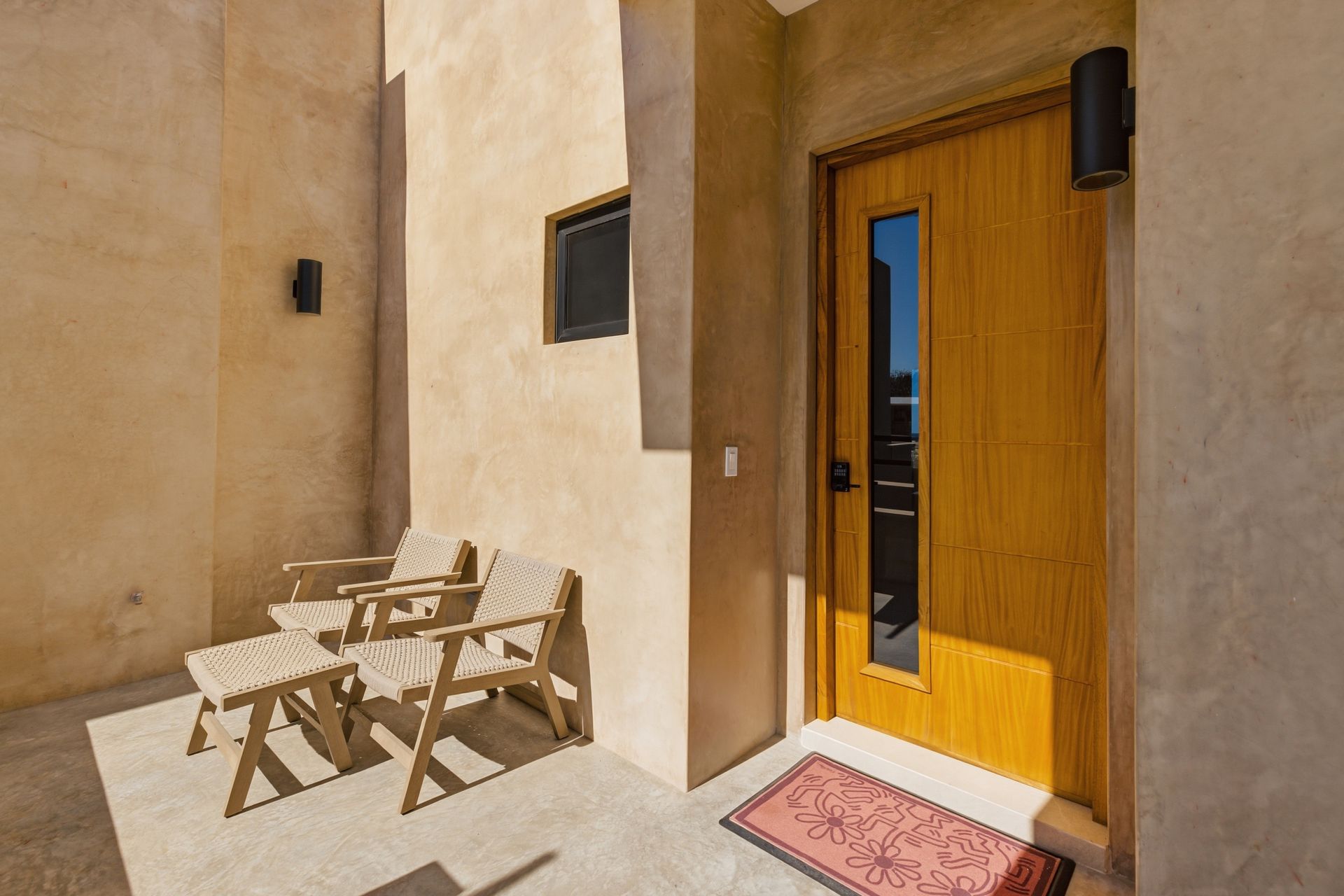 A wooden front door with a vertical glass panel, two patio chairs, and a small rug on a concrete porch against tan walls.