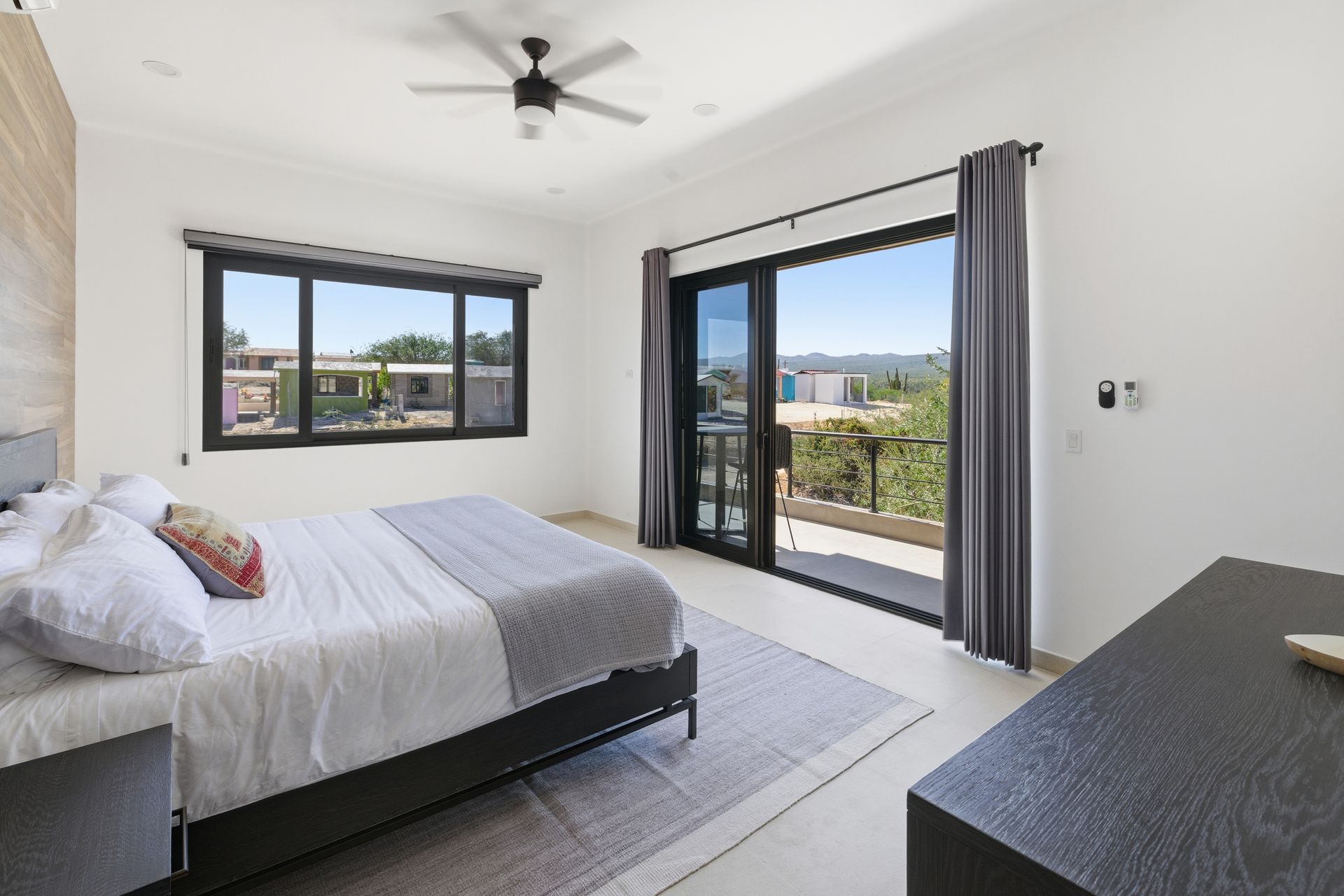 A bright bedroom featuring a queen bed with white bedding, a gray rug, a ceiling fan, and large windows leading to a deck.