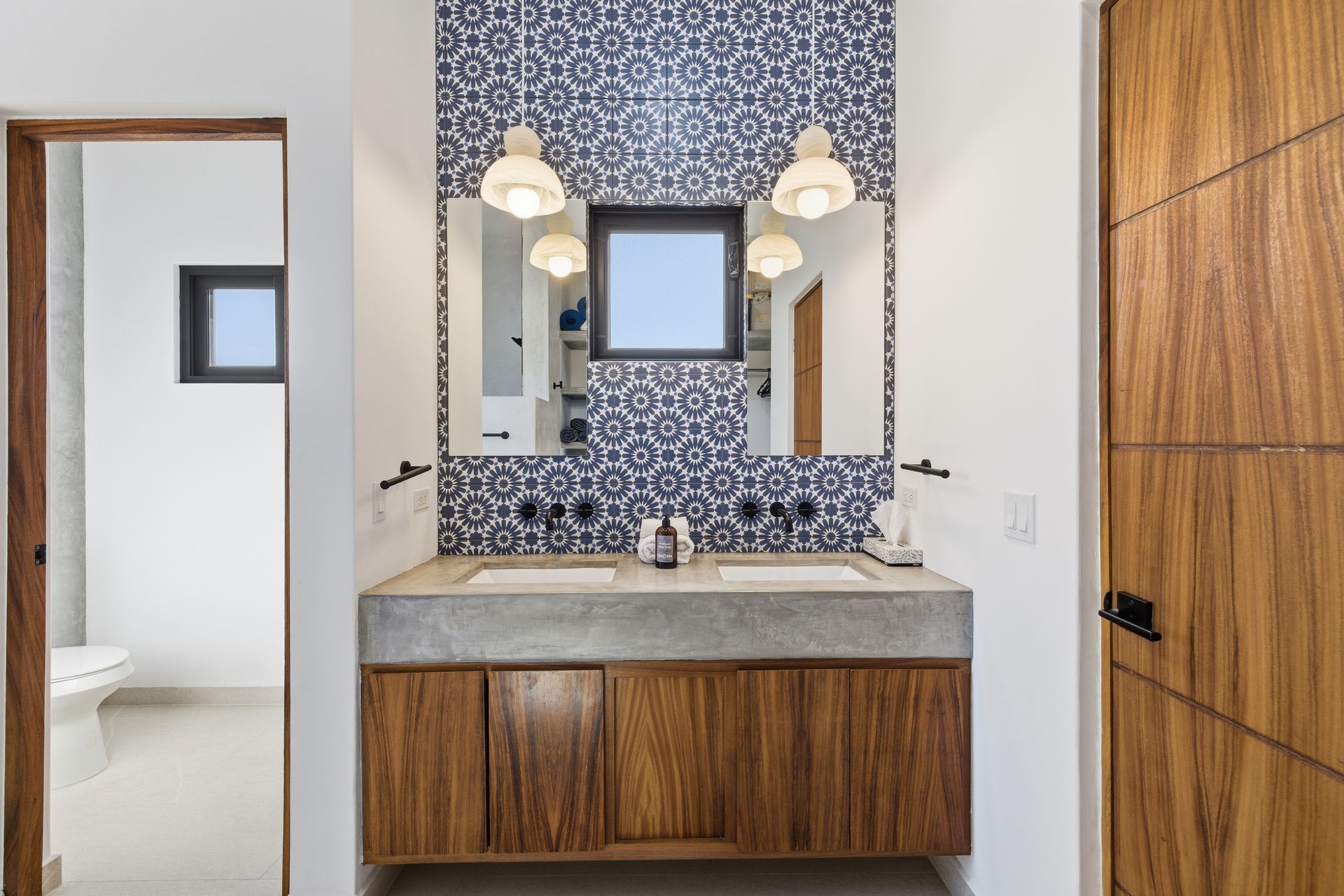 Modern bathroom with a double concrete vanity, blue patterned tile backsplash, wooden cabinets, and hanging globe lights.