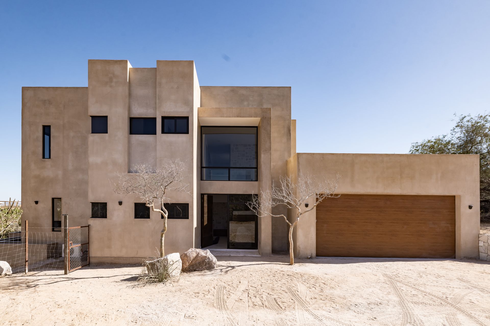 A modern, tan-colored two-story stucco house with a large wood garage door, set in a desert landscape under a clear sky.