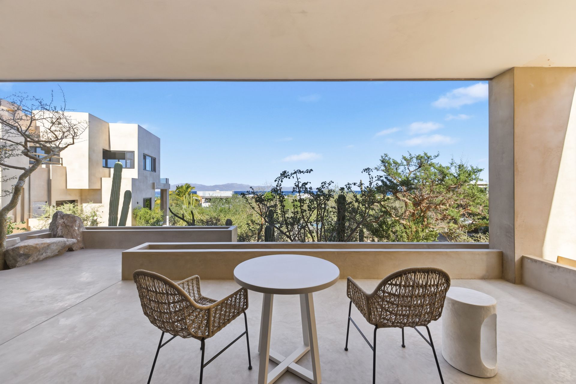 A covered outdoor terrace with two wicker chairs, a round table, and a desert view with distant mountains.