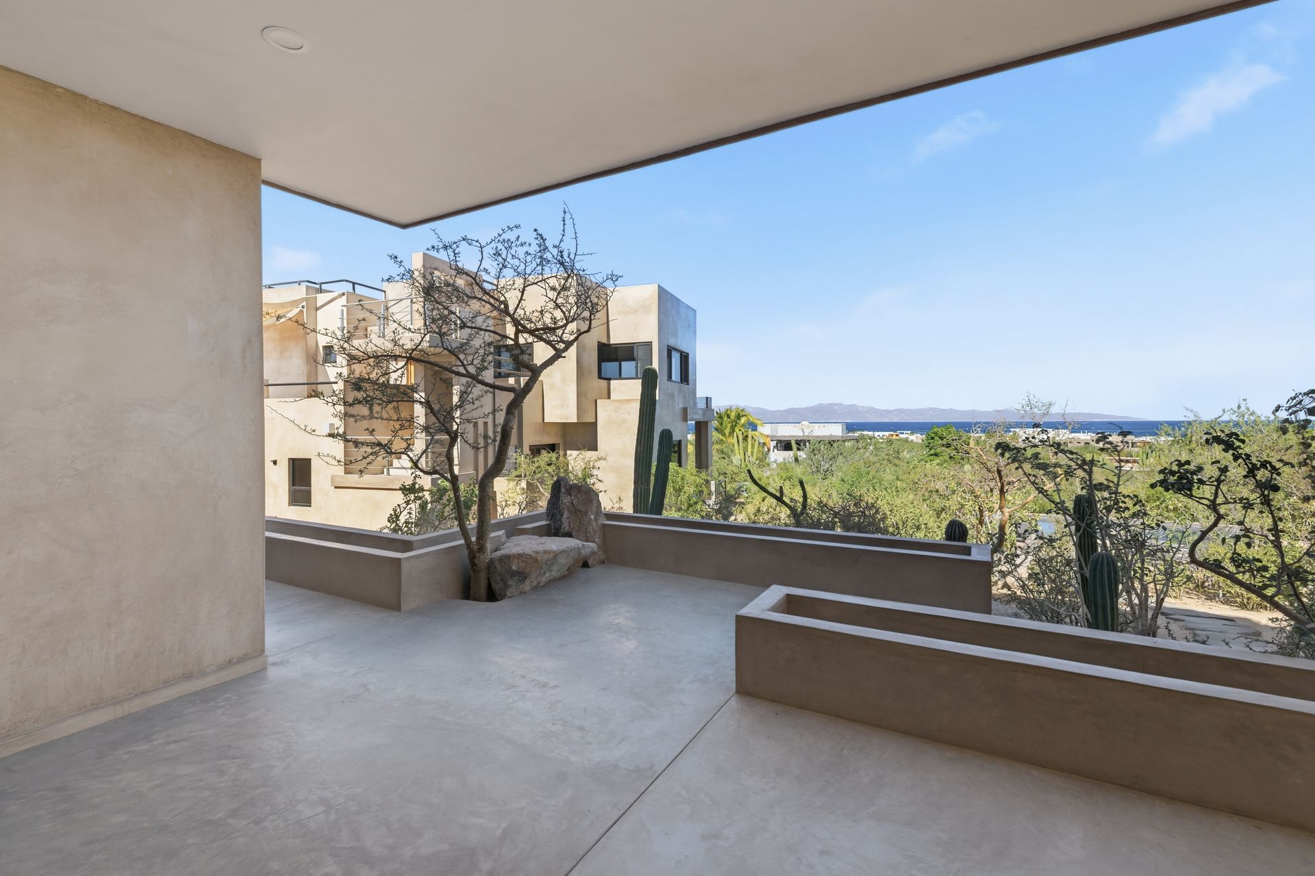 A modern concrete patio overlooks a desert landscape with a tree, cactus, and a distant blue ocean under a clear sky.
