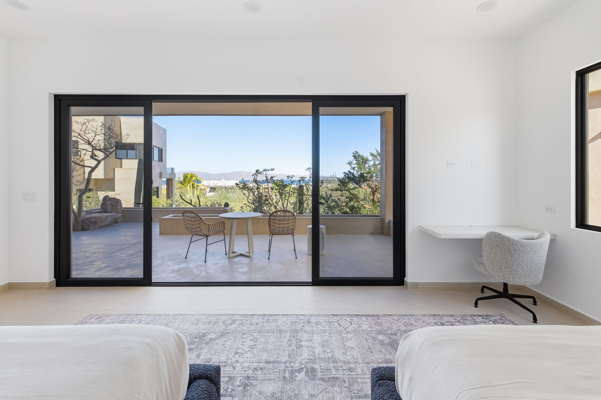 A bedroom with two beds, a white desk, and a large sliding glass door opening to a patio with outdoor furniture and a view.