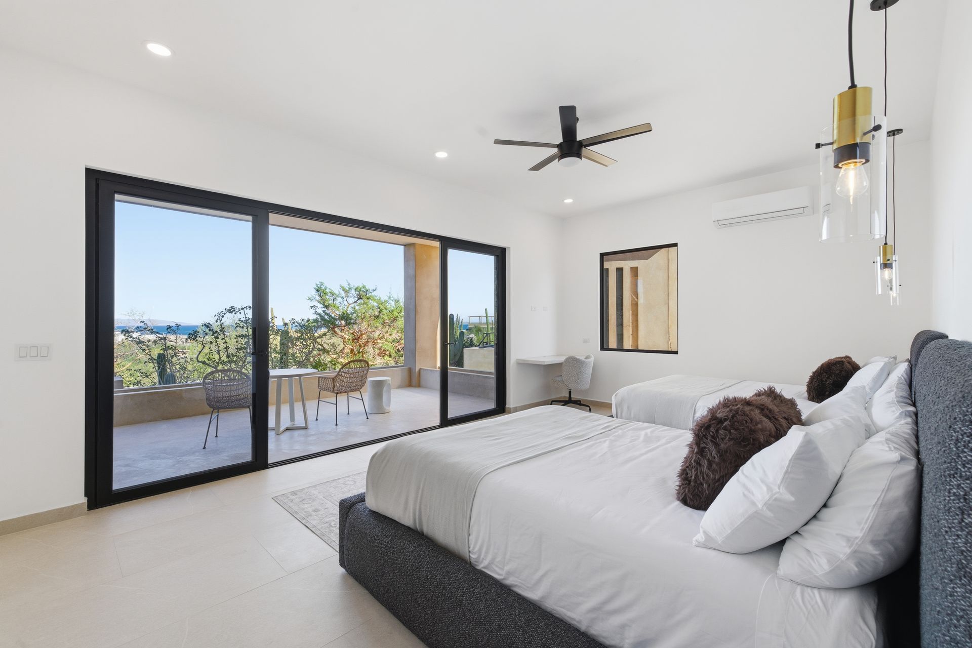 Modern bedroom with two beds, white walls, a ceiling fan, and large sliding doors opening to a sunny balcony.