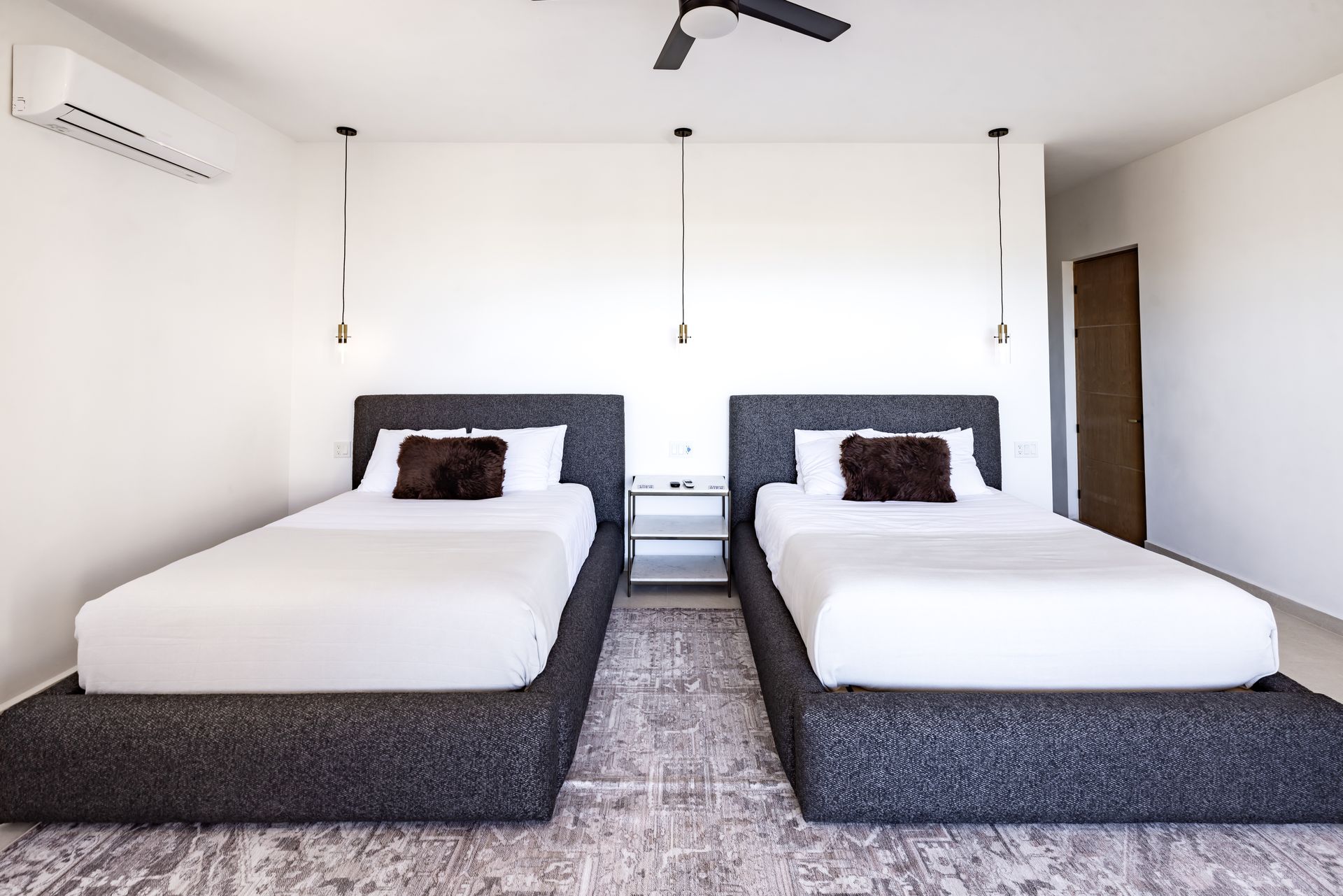 Two beds with dark gray upholstered frames, white bedding, and small brown pillows in a minimalist, white-walled room.