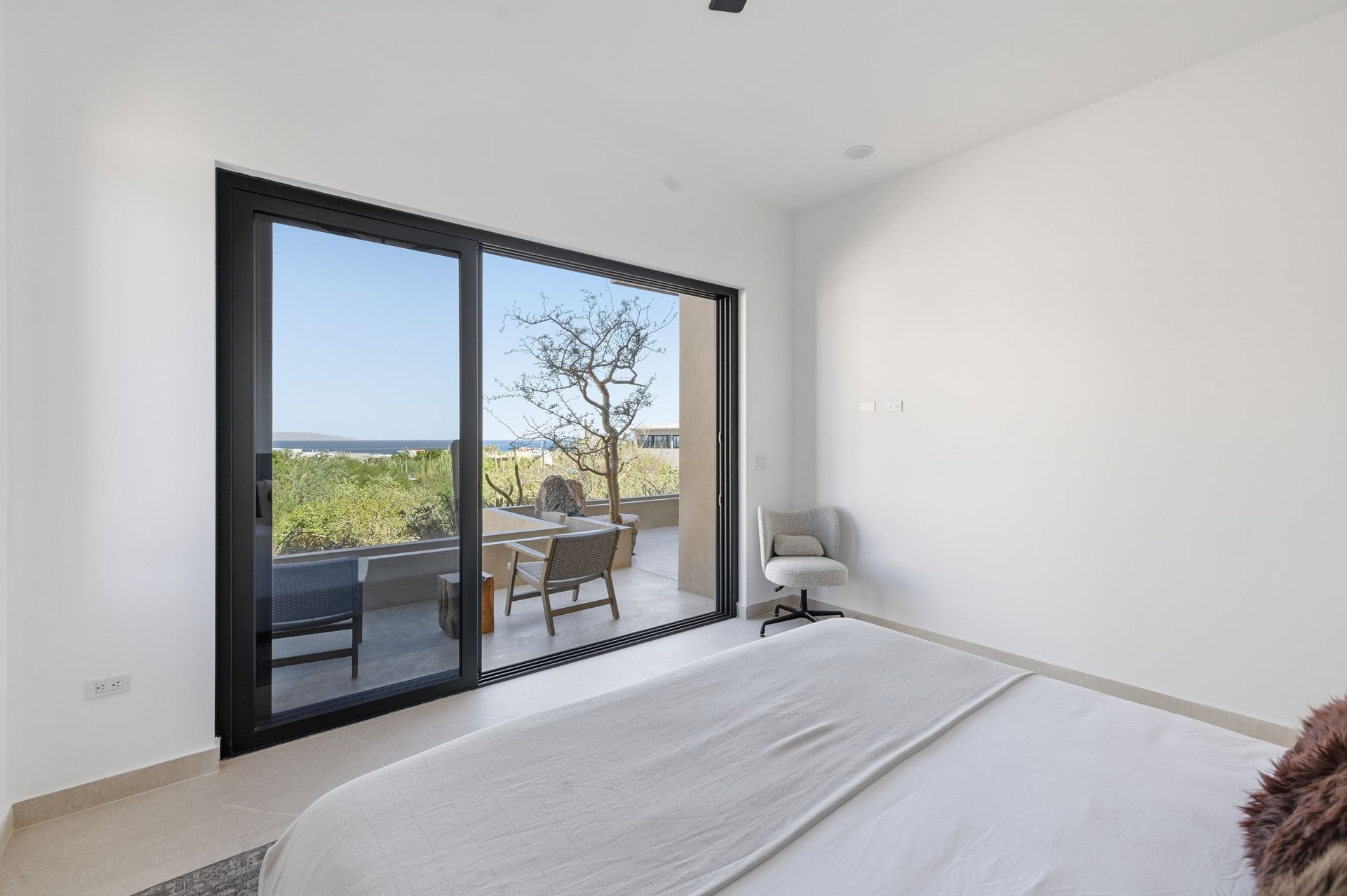 Modern bedroom with a large sliding glass door opening to a patio featuring an outdoor chair and coastal view.
