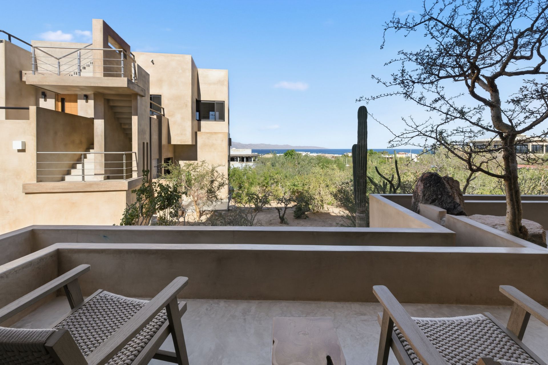 A tan stucco balcony with two lounge chairs overlooking a desert landscape with cacti and a distant sea view.