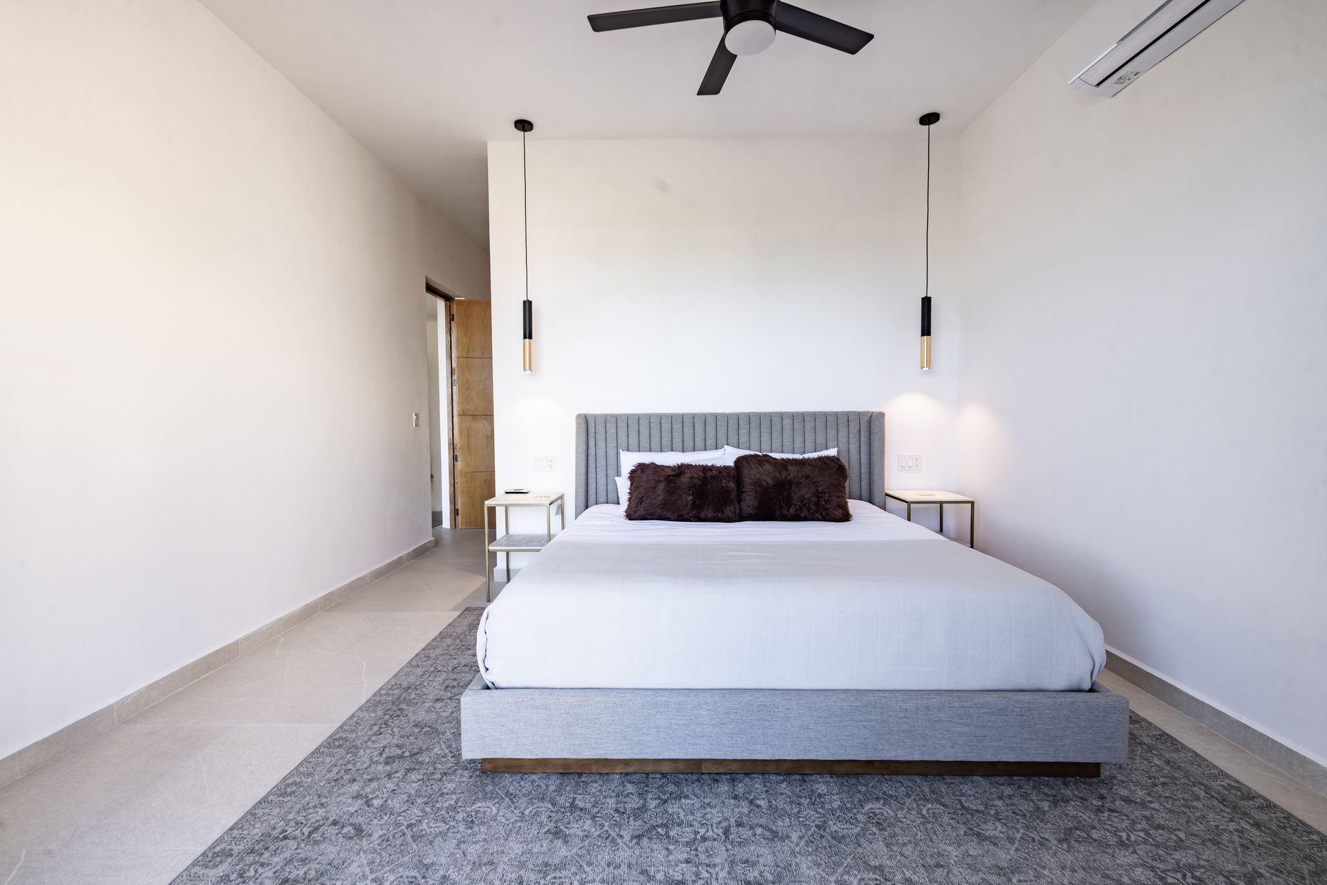 A minimalist bedroom featuring a gray upholstered bed, two hanging pendant lights, and a gray area rug on a tile floor.