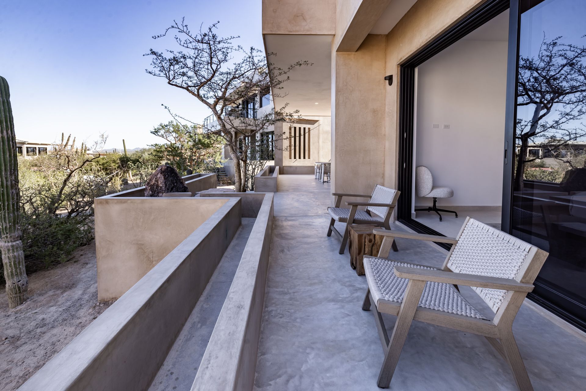 A modern balcony with woven chairs overlooking a desert landscape with cacti and sparse trees under a clear blue sky.