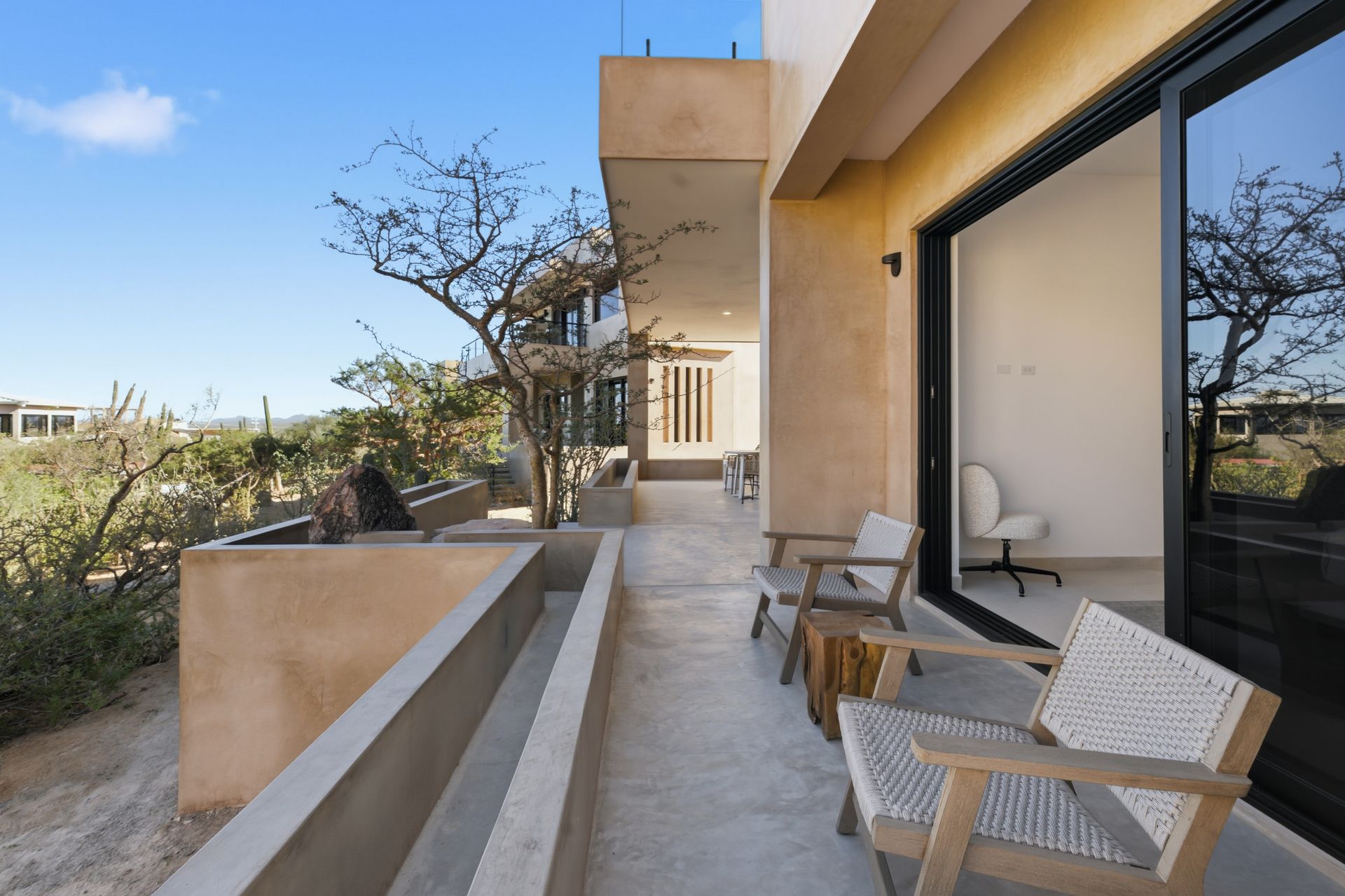 A sunlit balcony with woven chairs overlooking a natural, desert-like landscape under a clear blue sky.