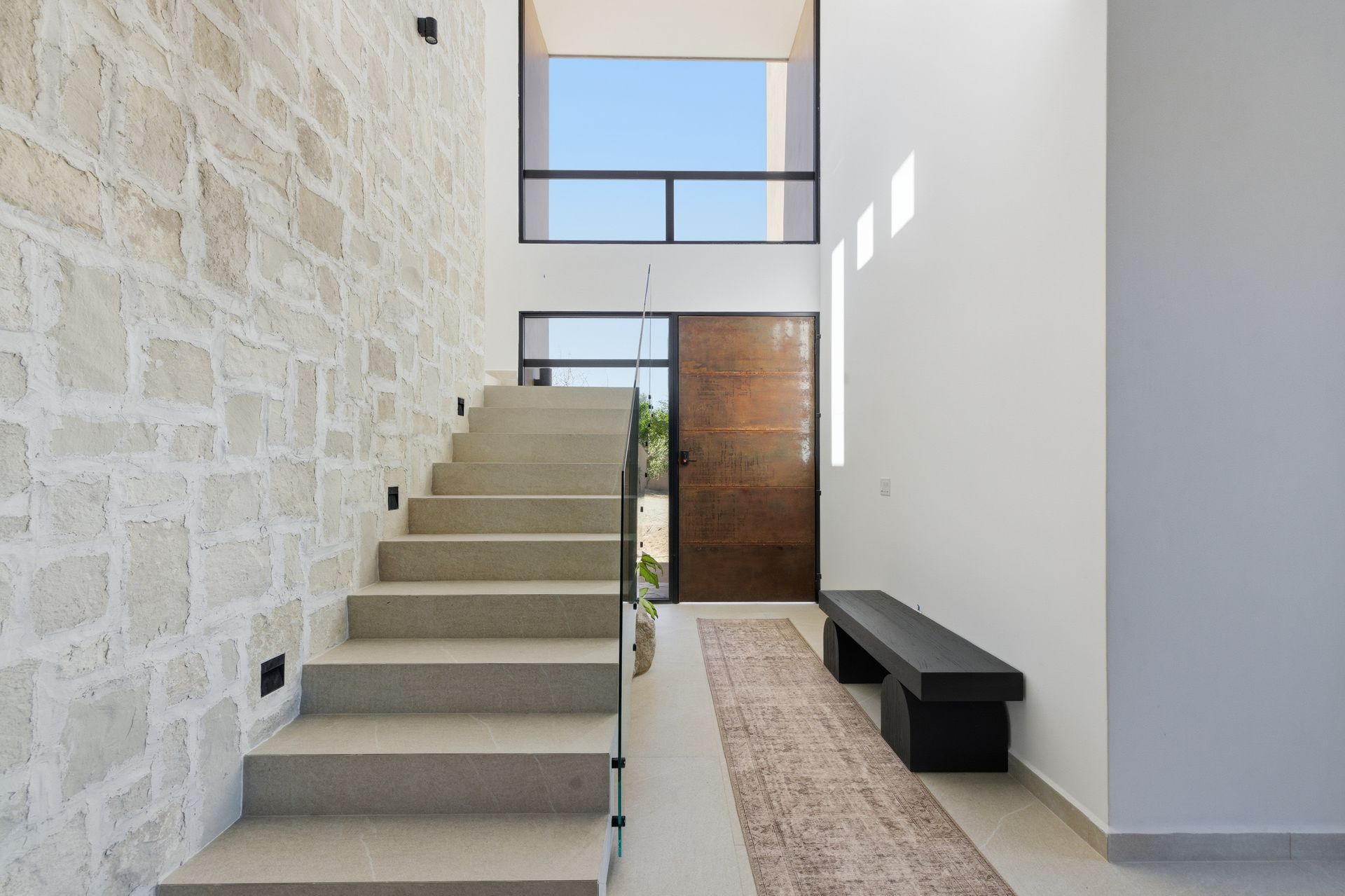 A bright entryway featuring a stone wall, a floating concrete staircase, a dark wood front door, and a black bench.