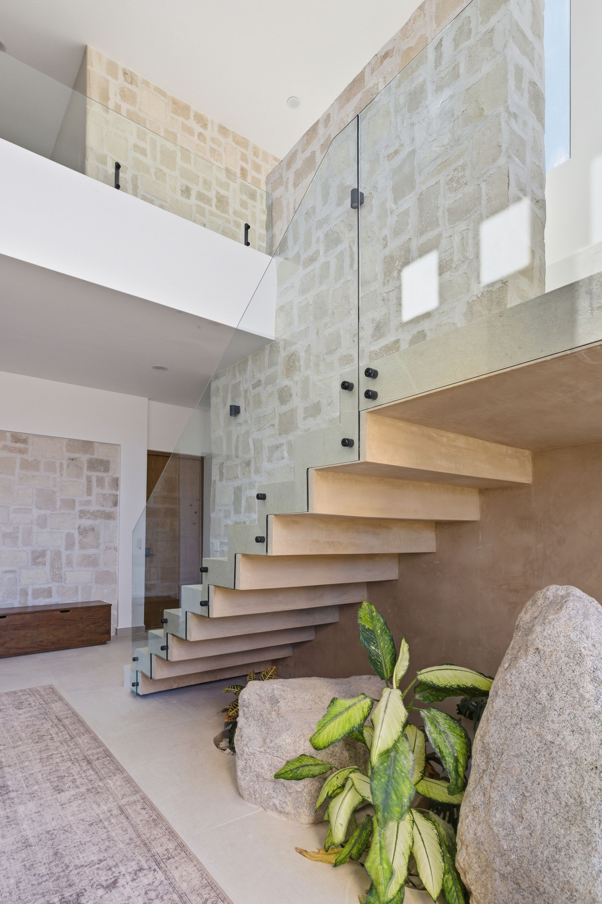Modern floating wooden staircase beside a stone wall with a potted plant and large decorative stones in an open interior.