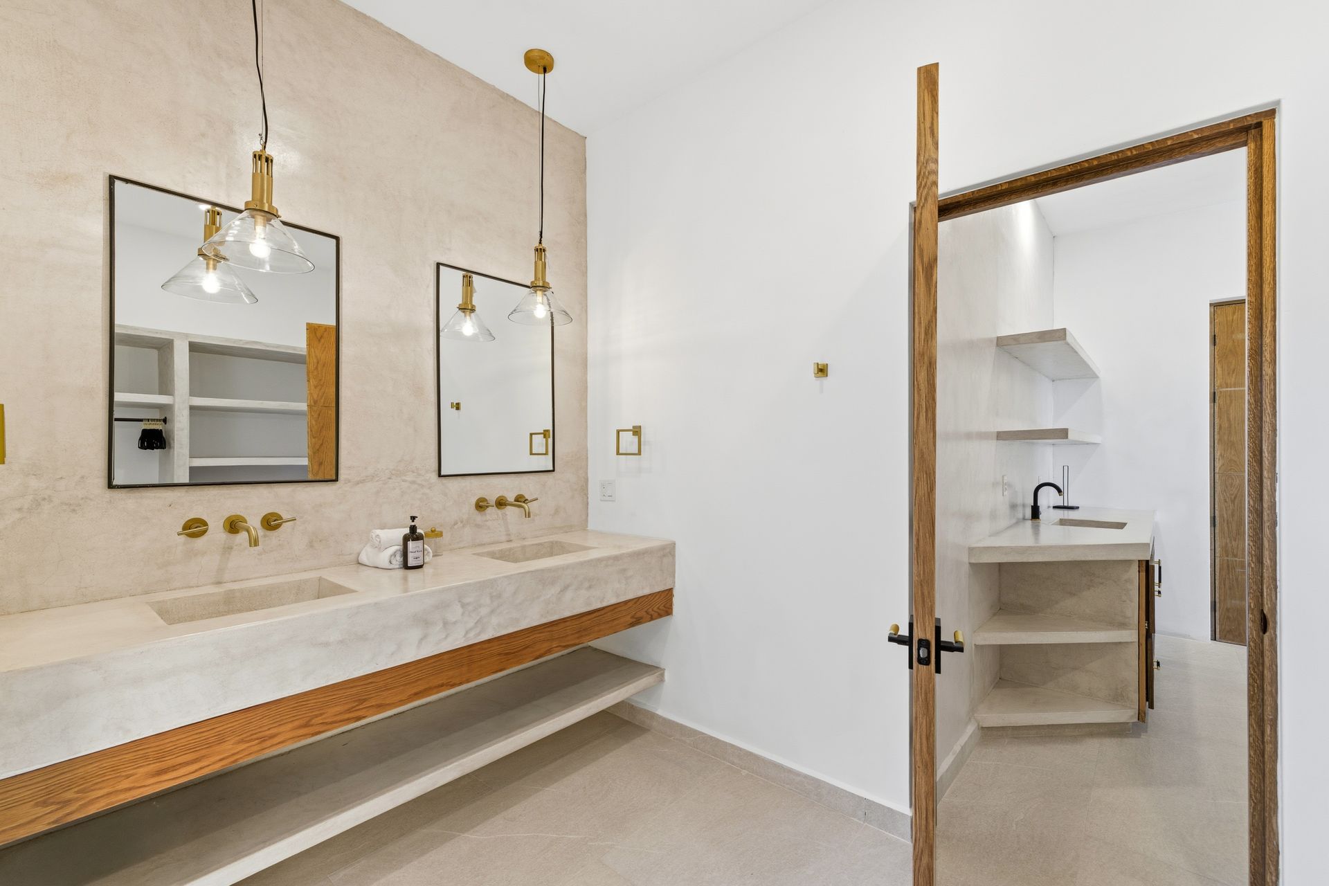 A minimalist bathroom with a double stone vanity, gold fixtures, two pendant lights, and a doorway to a small kitchenette.