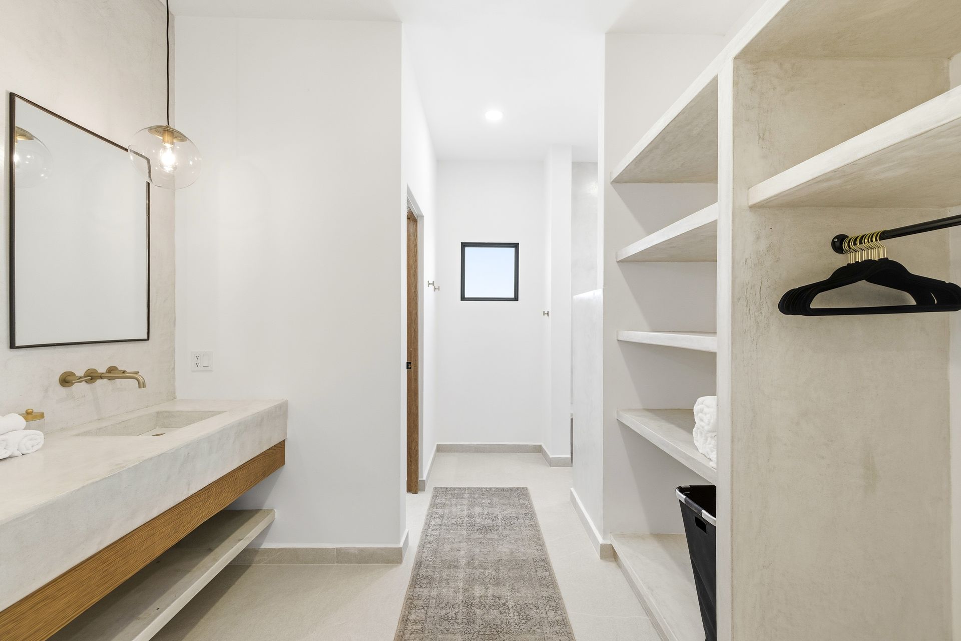 A bright, modern bathroom with a stone sink, minimalist shelves, a black hanger, and a runner rug leading to a hallway.