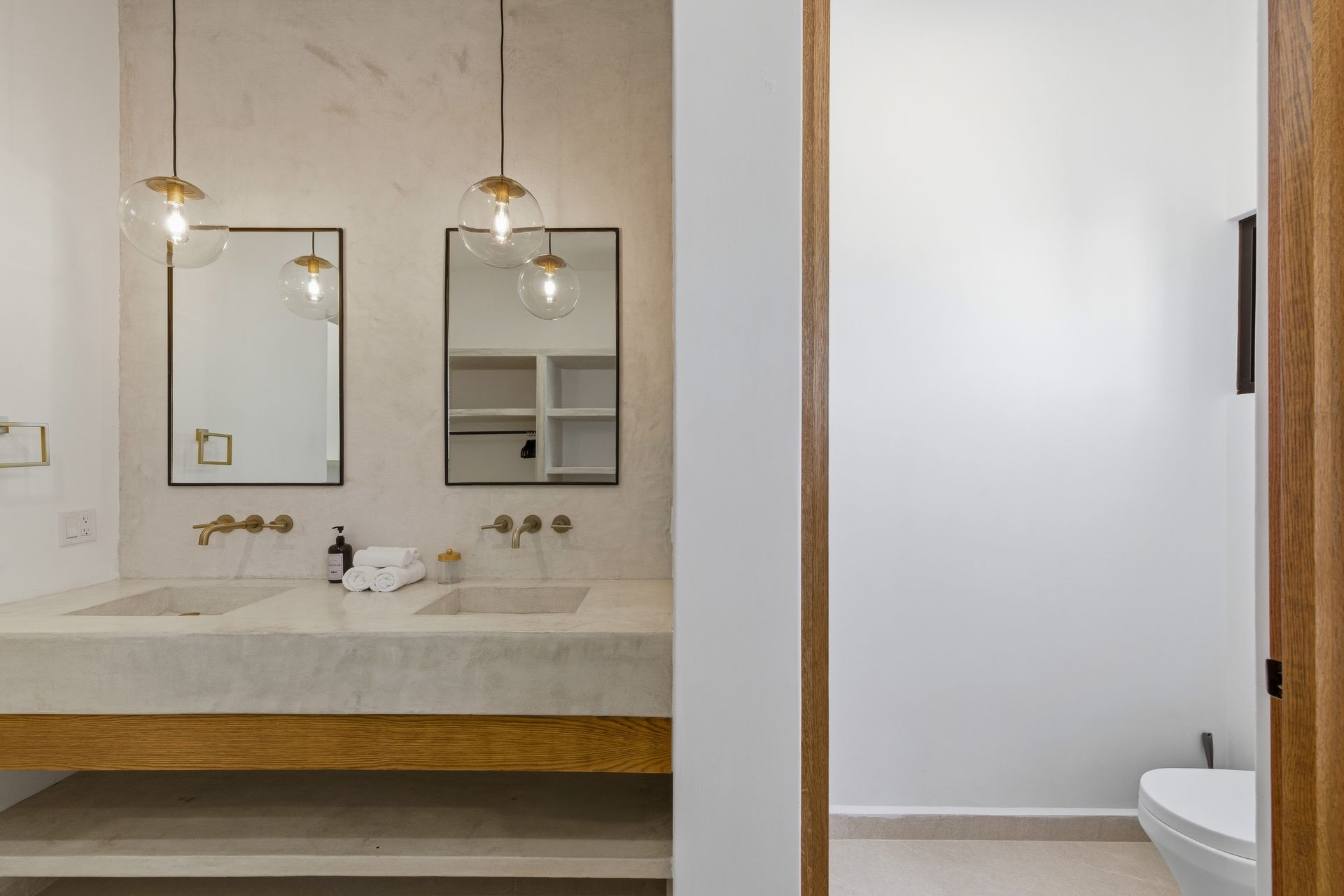 A modern bathroom with a double stone vanity, brass fixtures, two hanging pendant lights, and an adjacent toilet area.