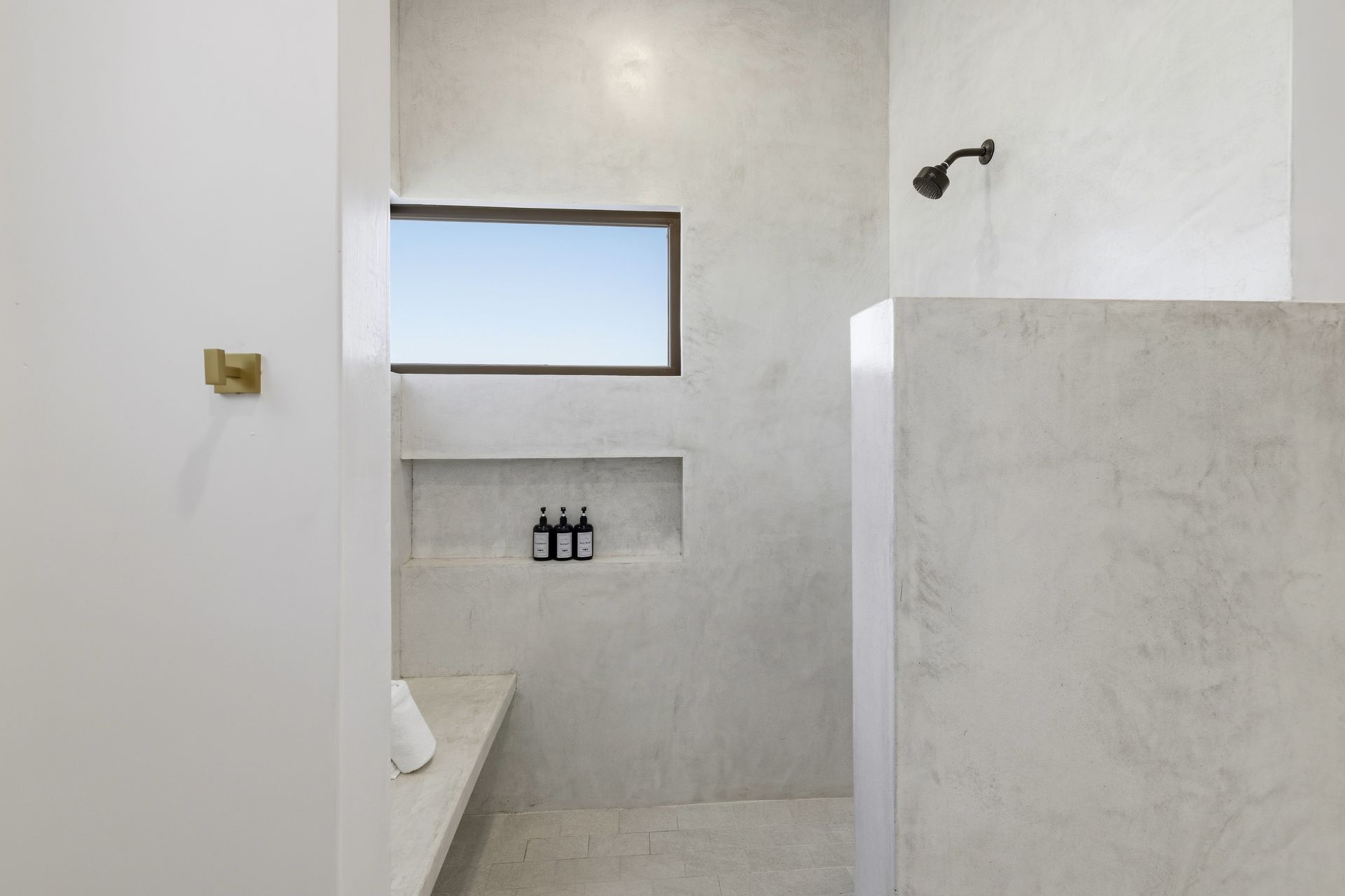 A minimalist walk-in shower with light gray concrete walls, a small window, built-in shelving, and a black showerhead.