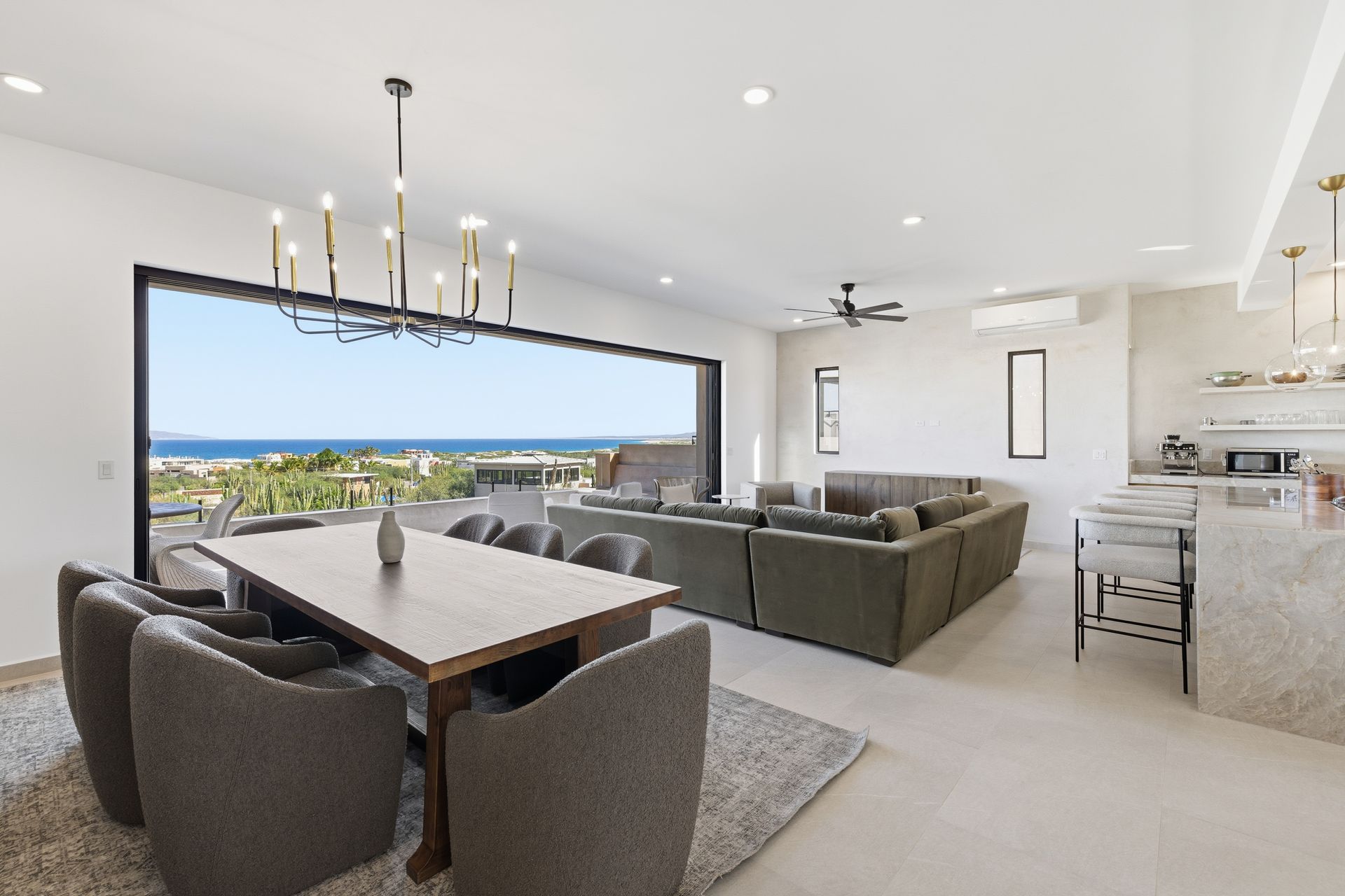 Open-plan living room and dining area with modern decor, a large sofa, and a scenic coastal view through sliding doors.