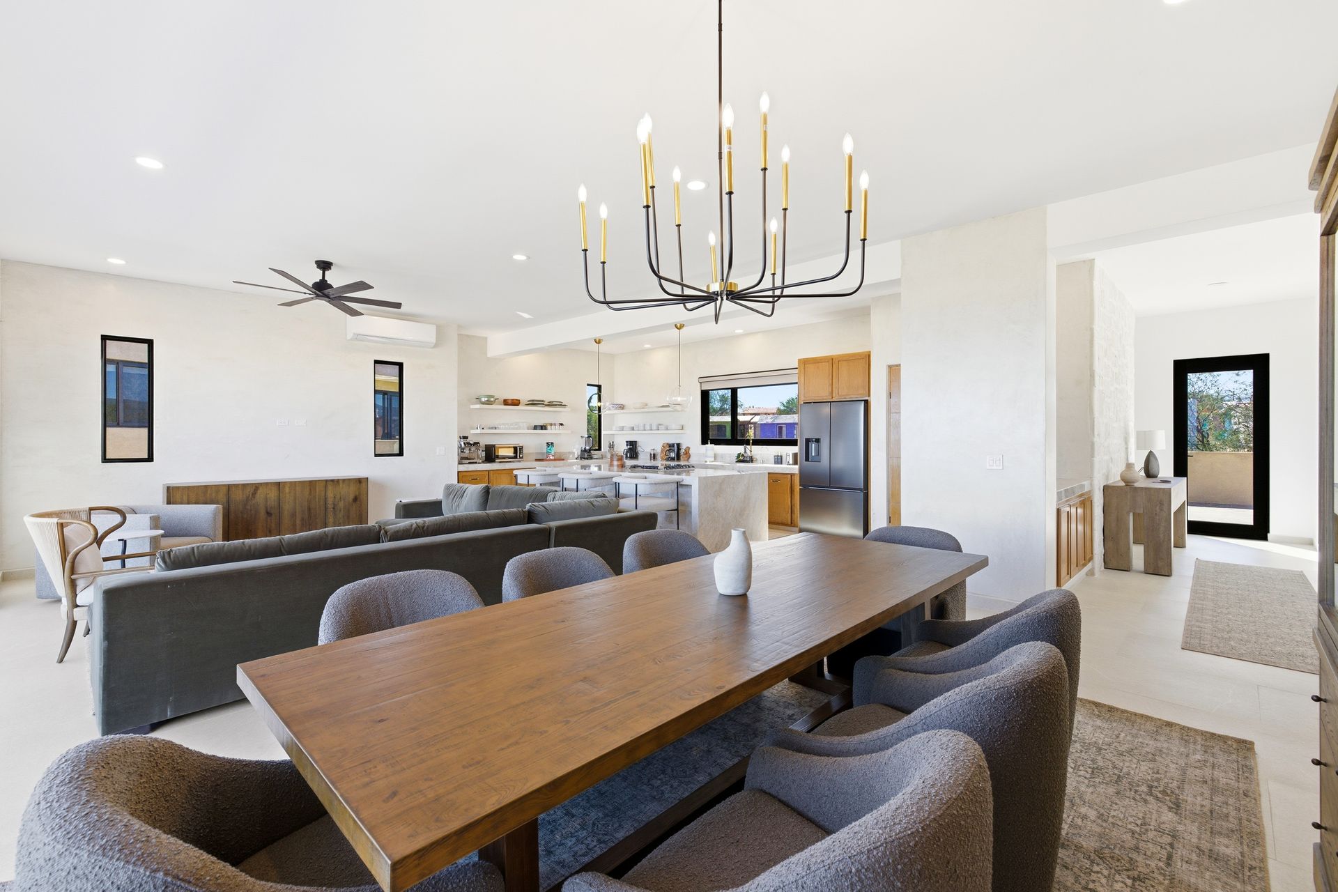 An open-concept dining and living area with a long wooden table, grey chairs, a modern chandelier, and a kitchen background.