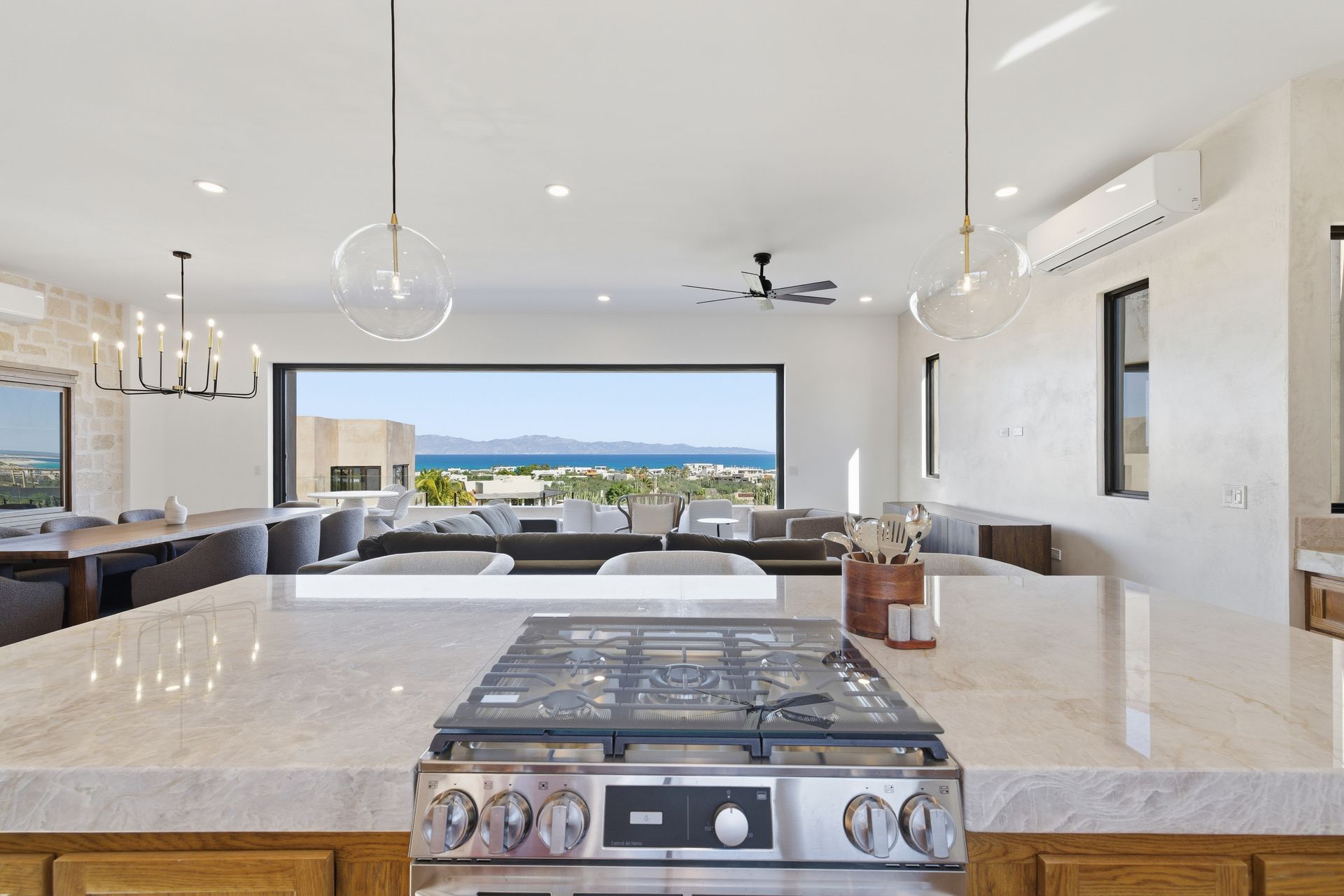 A modern kitchen island with a gas stovetop overlooks an open-concept living area with large windows showing a coastal view.