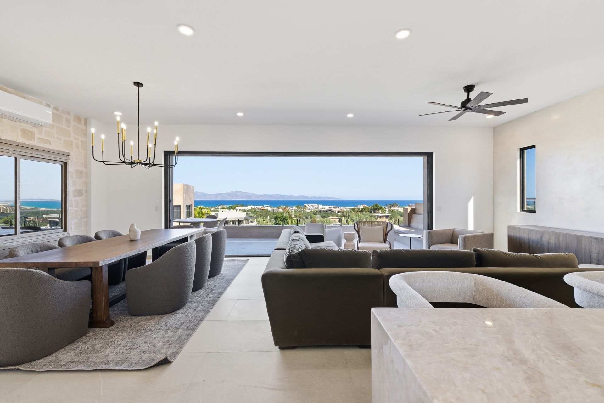 A modern, open-concept living and dining room with large windows overlooking a coastal view and mountains.