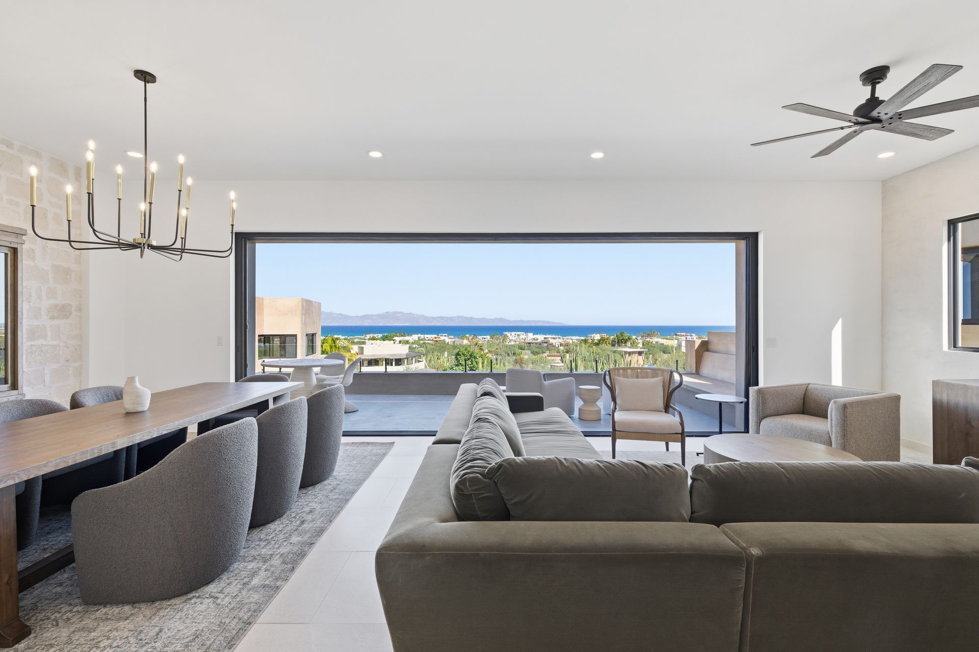Modern living room with a long dining table, grey armchairs, large sectional sofa, and a scenic ocean view through glass.