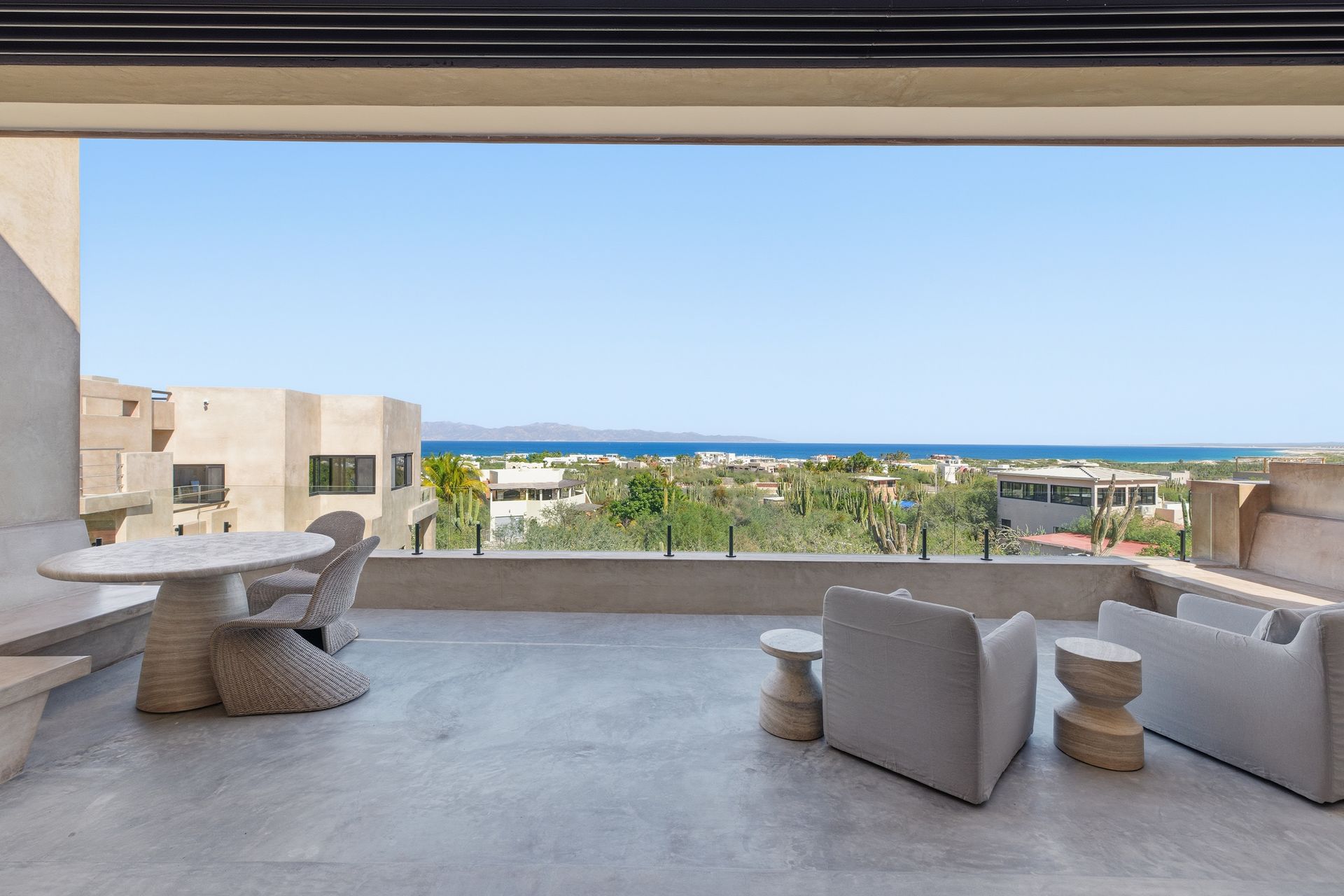 A modern outdoor patio with white dining furniture and lounge seating, overlooking a lush garden and the blue ocean.