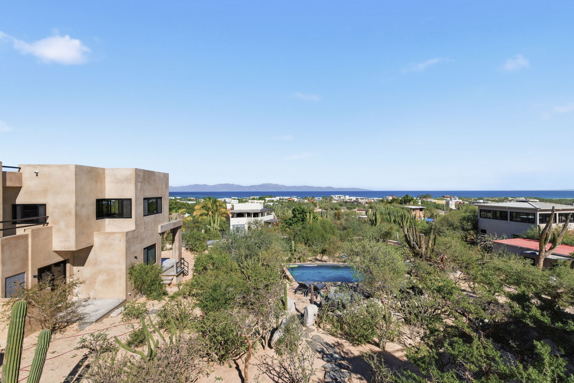 A tan, multi-level house overlooks a landscape of desert scrub, cacti, and a small pool, with the ocean and hills beyond.