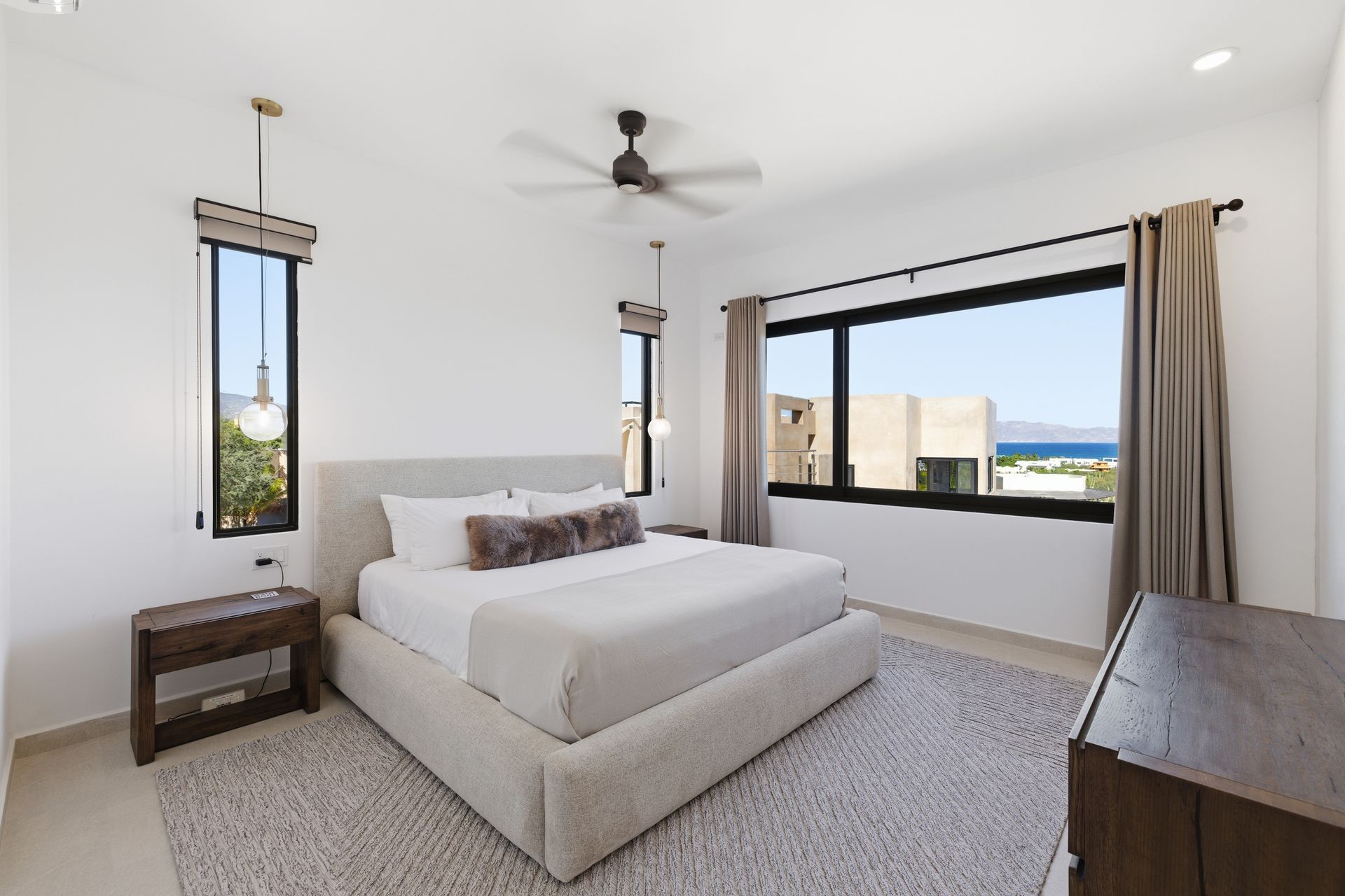 A bright, modern bedroom with a large upholstered bed, wooden furniture, and a window showcasing a scenic coastal view.