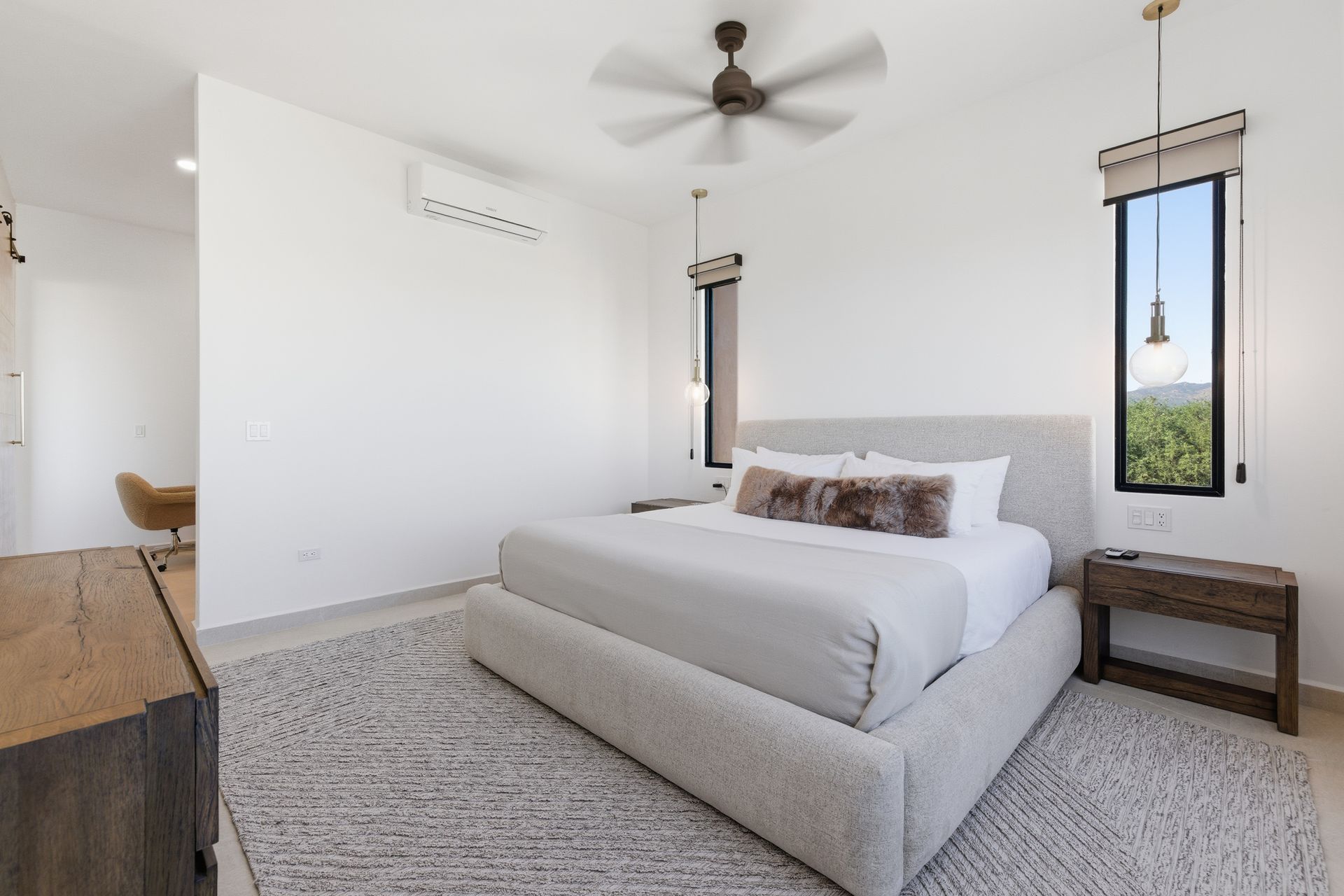 A modern bedroom with a gray upholstered bed, light rug, wood side table, minimalist light fixtures, and white walls.