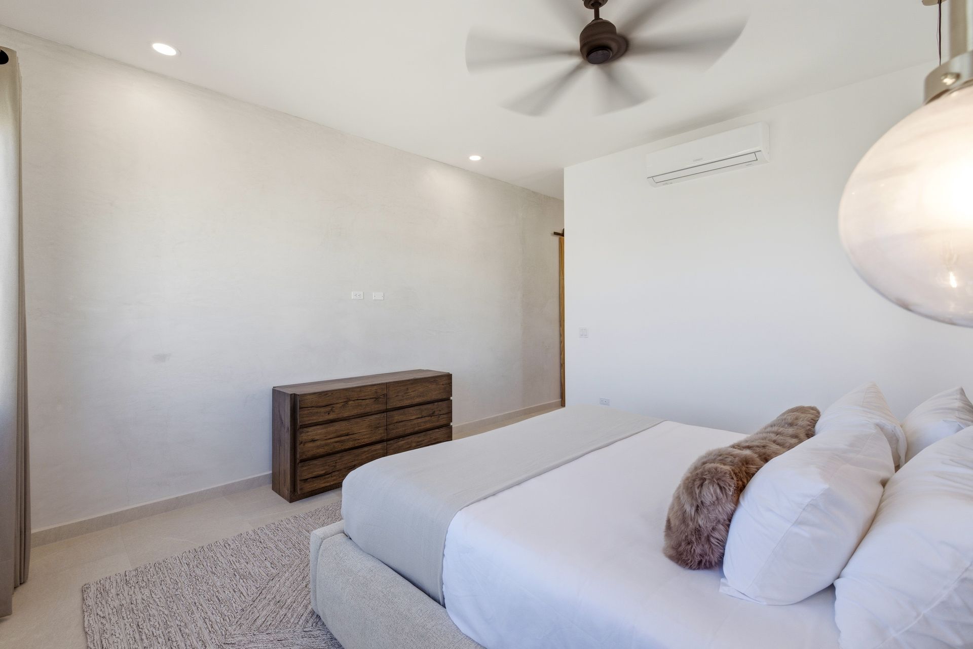 A bright, modern bedroom with a large bed, a wooden dresser, a ceiling fan, and a round pendant light.
