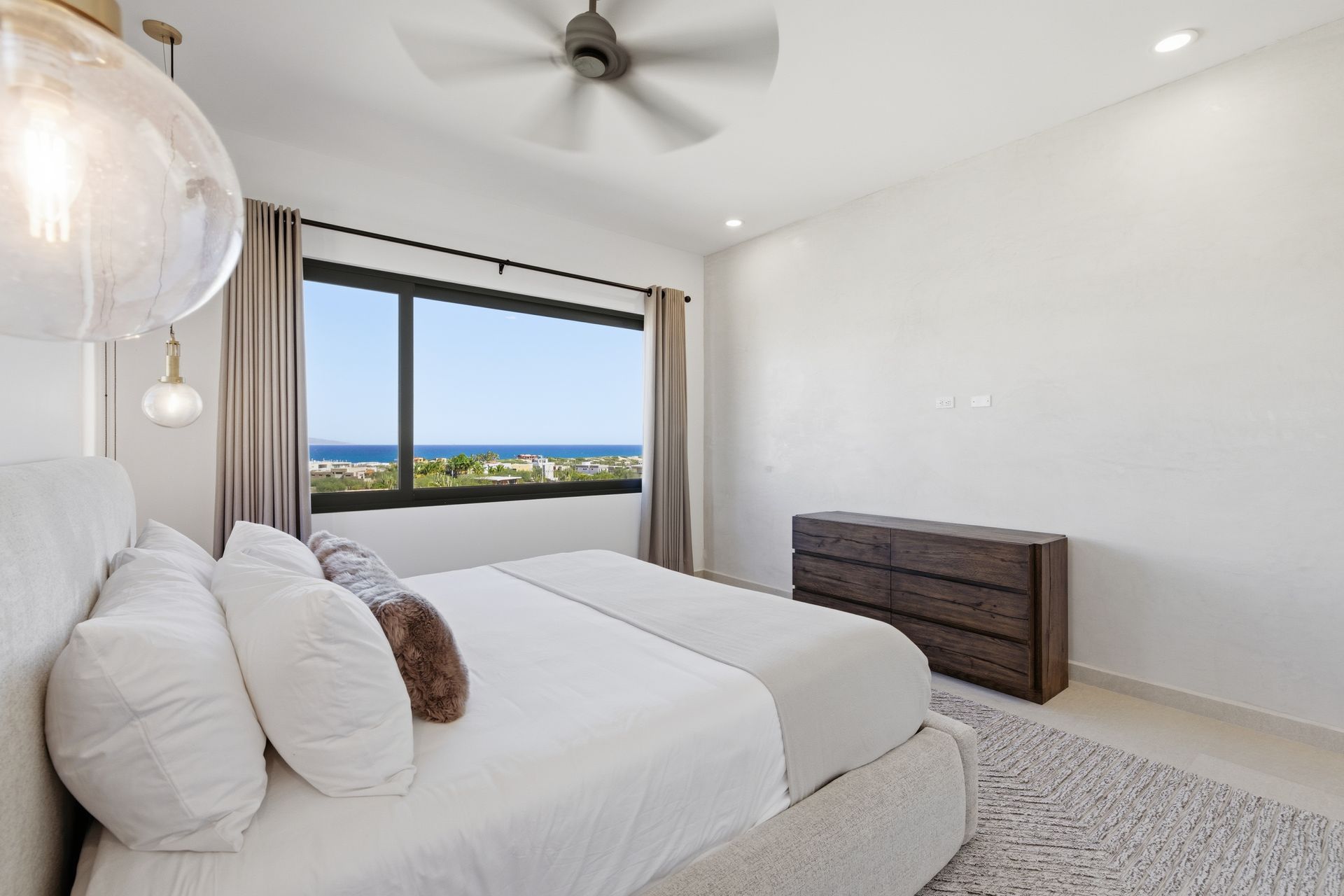 A bright bedroom featuring a queen bed with white linens, a wooden dresser, and a large window overlooking the ocean.