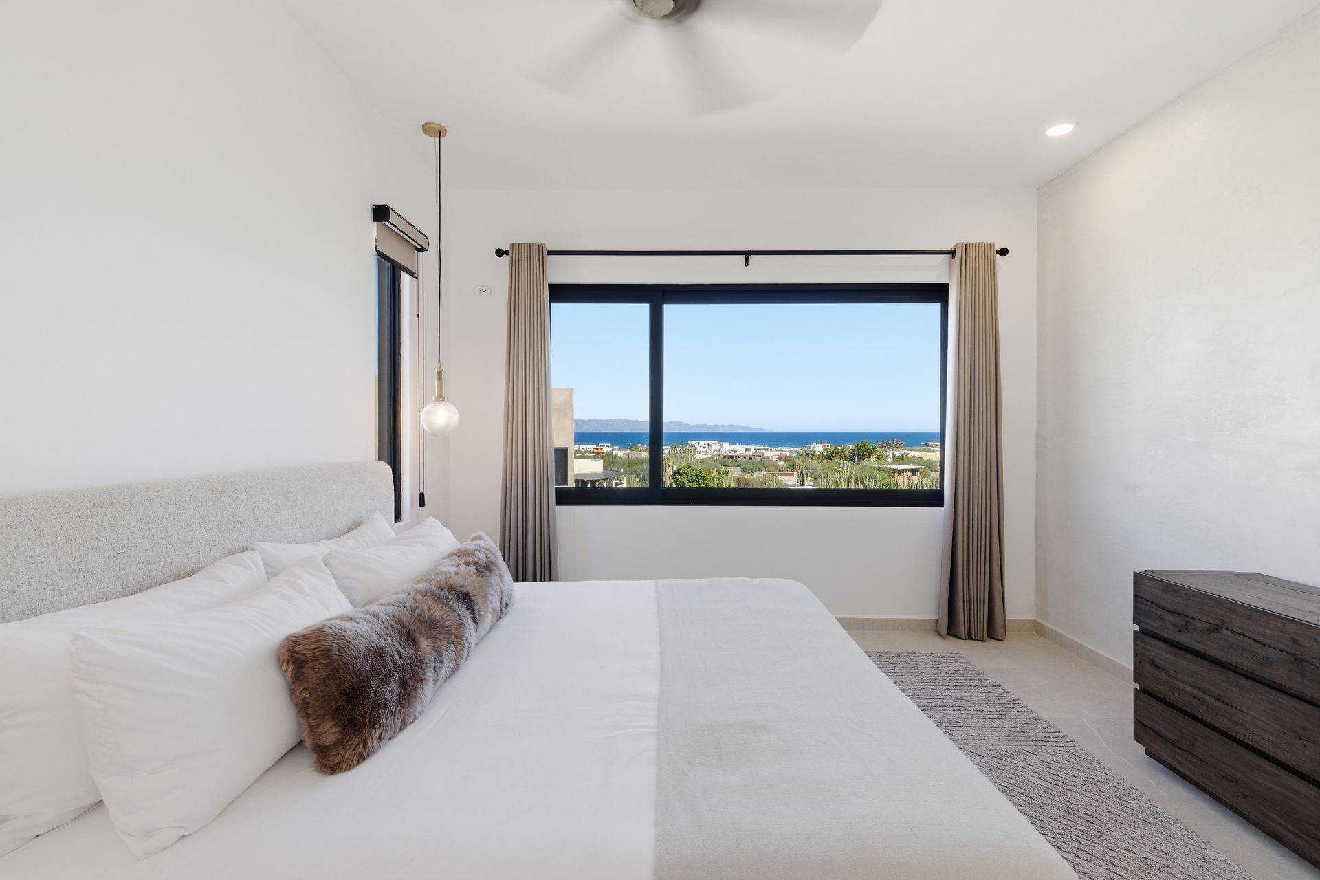 A bright modern bedroom with a large bed, brown bolster pillow, and a wide window offering a scenic ocean view.