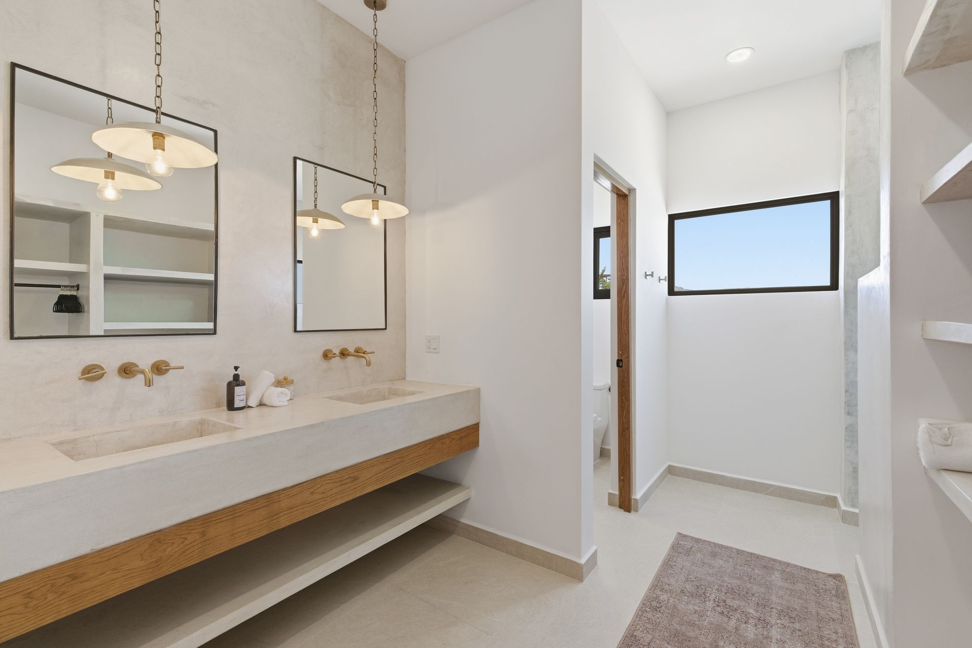 A modern bathroom featuring a double vanity, gold faucets, mirrors with hanging lights, and a window to a tiled shower.