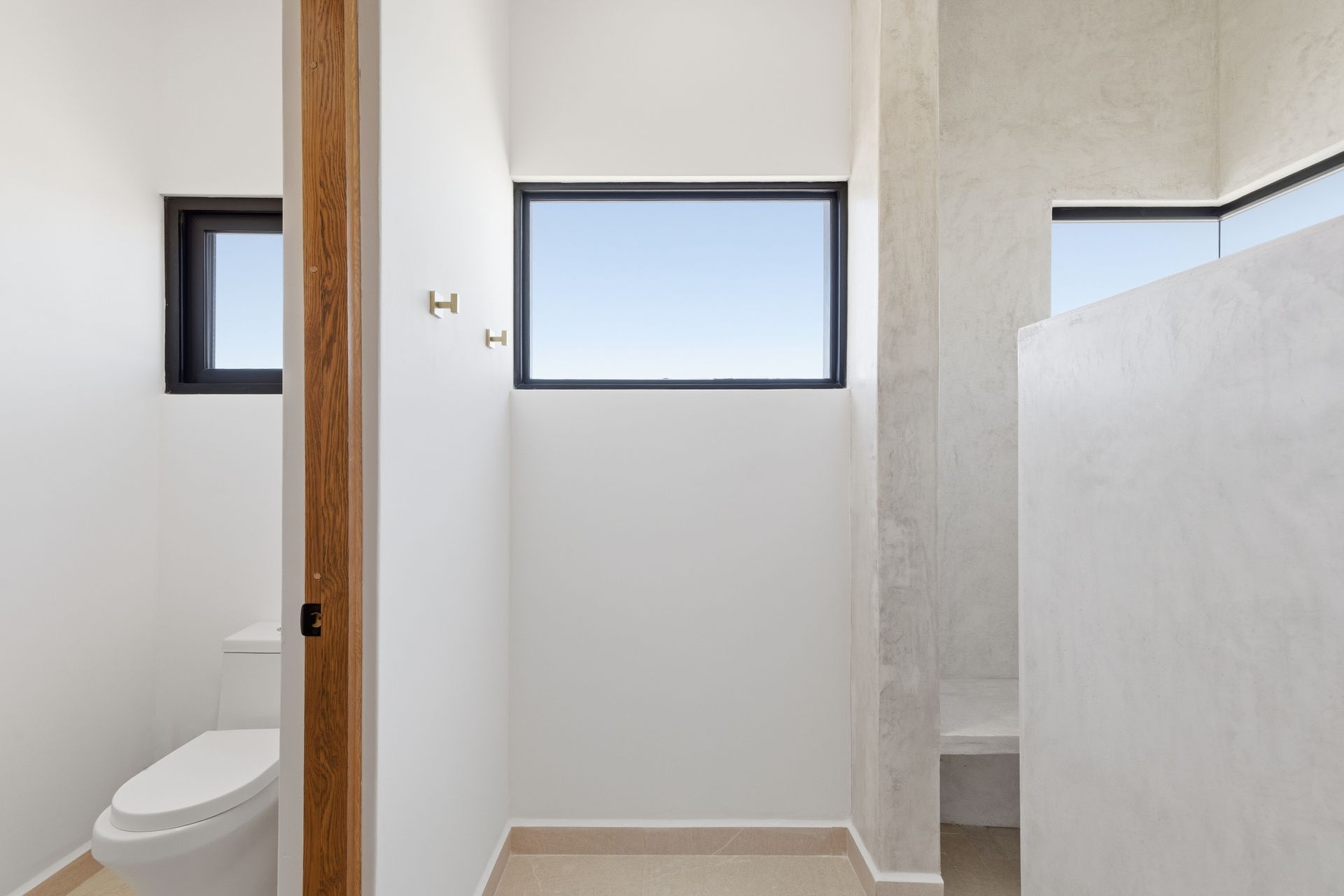 A modern, minimalist bathroom with white walls, two horizontal windows, a wooden door frame, and a white toilet.