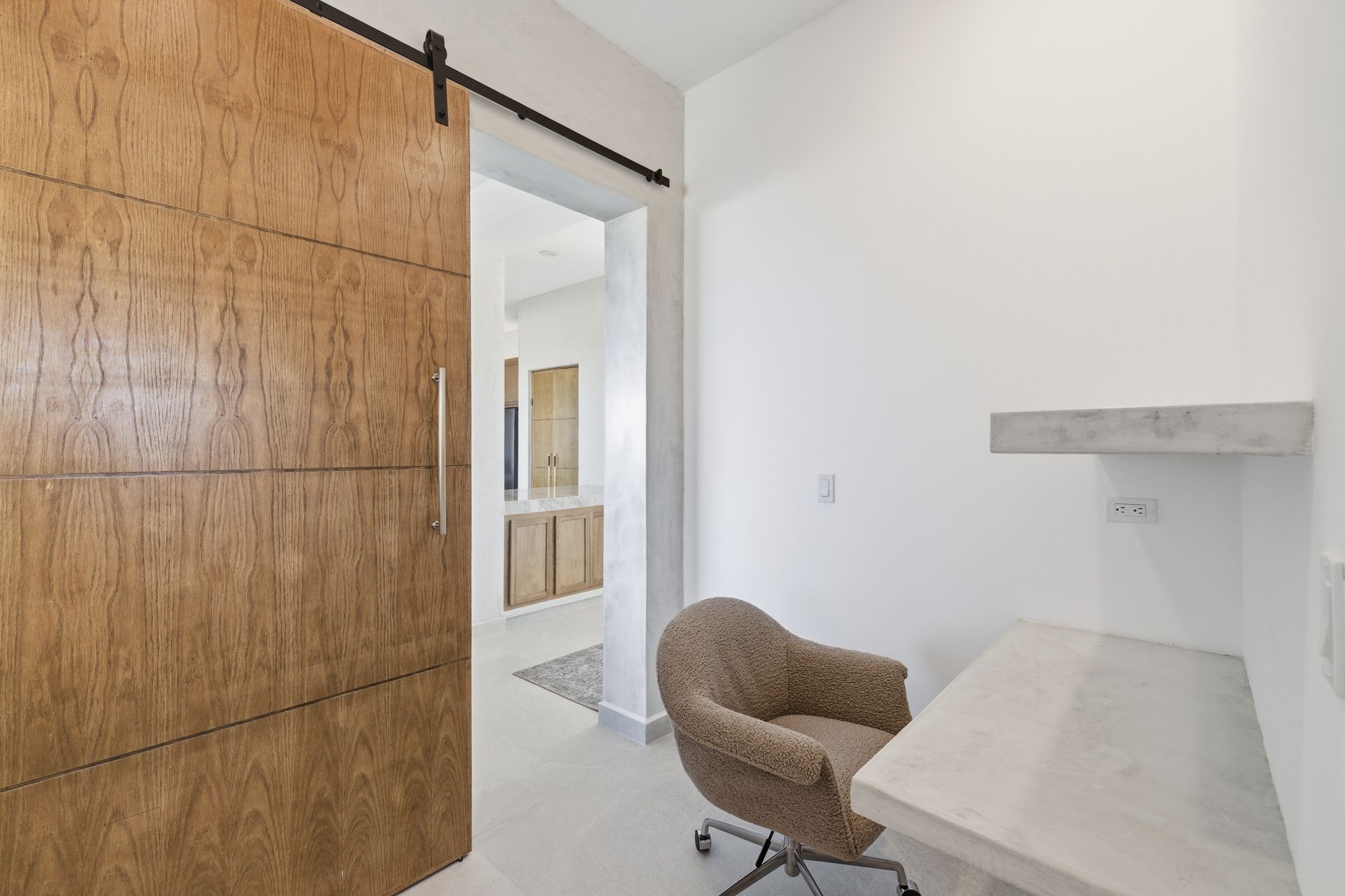 A home office nook with a textured wooden barn door, a minimalist concrete desk, and a cushioned armchair.