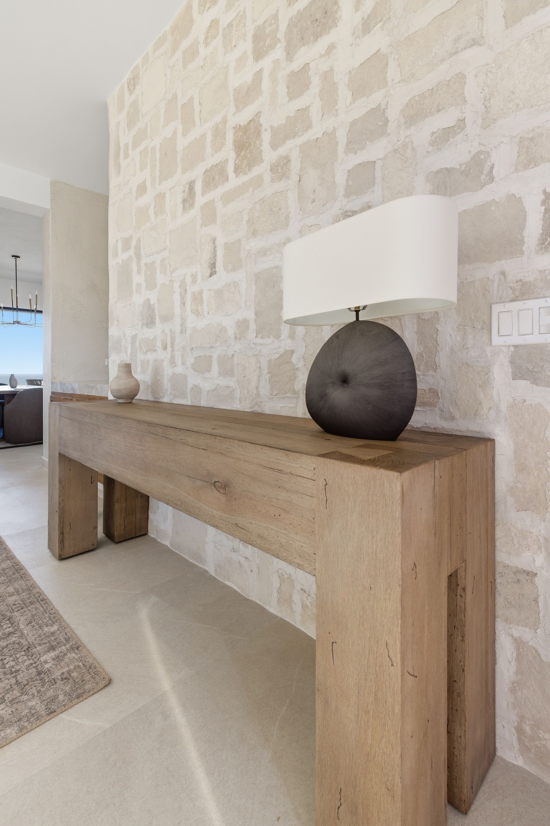 A rustic wooden console table with a dark stone lamp against a light-colored stone wall in a modern home interior.