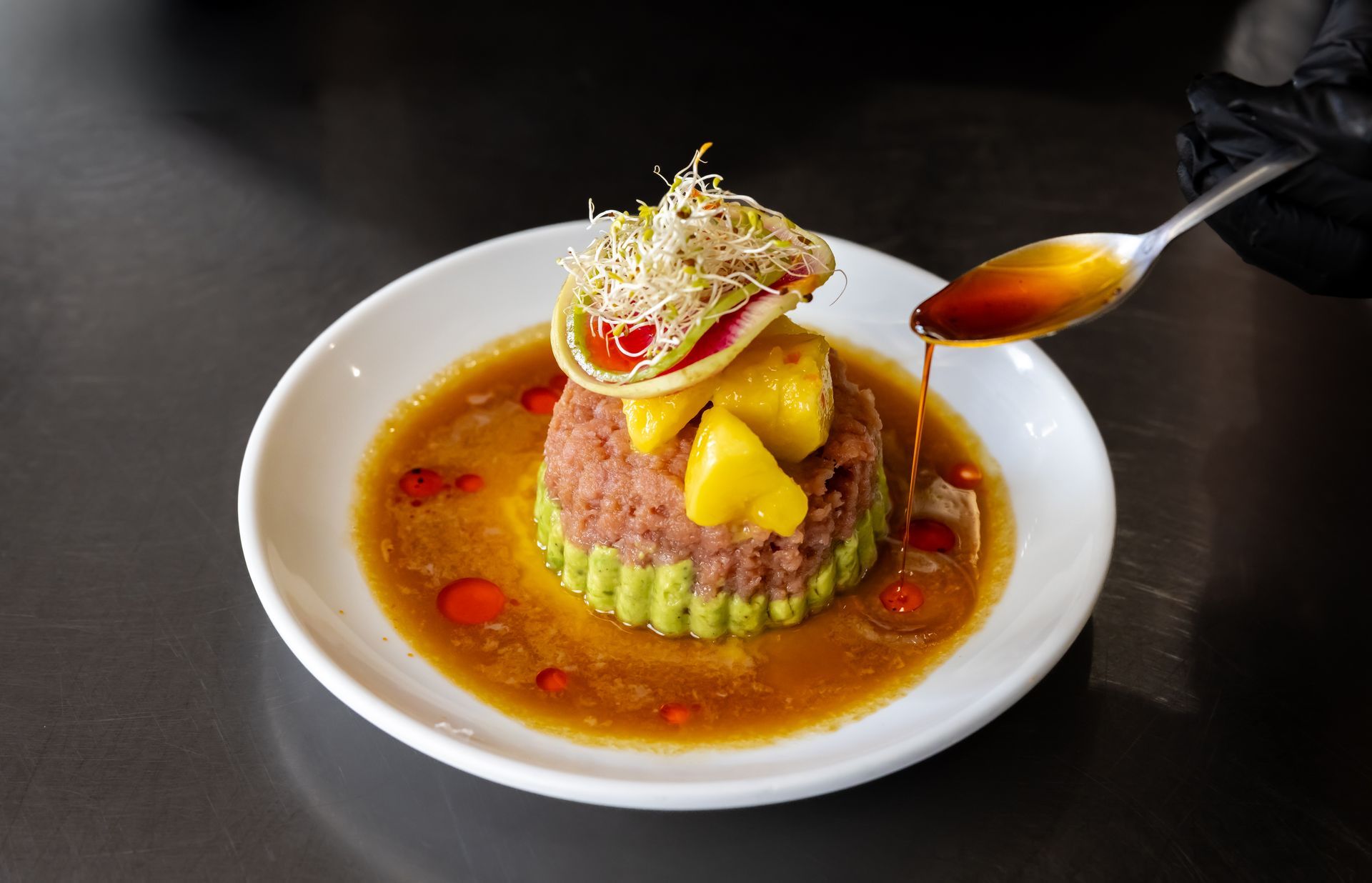 A gloved hand pours sauce from a spoon over a molded tuna and avocado tartare dish garnished with sprouts and mango.