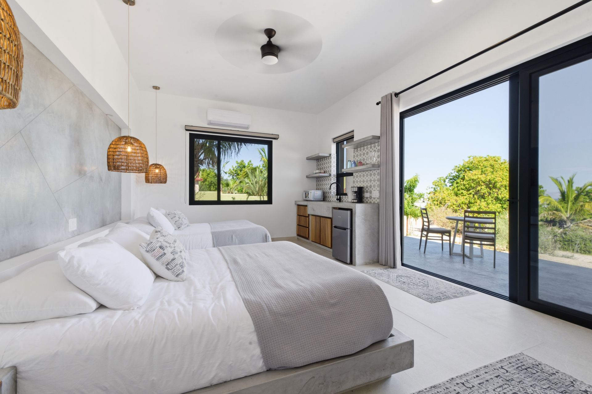 Modern bedroom with two beds, woven pendant lights, and large glass sliding doors opening onto a patio with outdoor chairs.