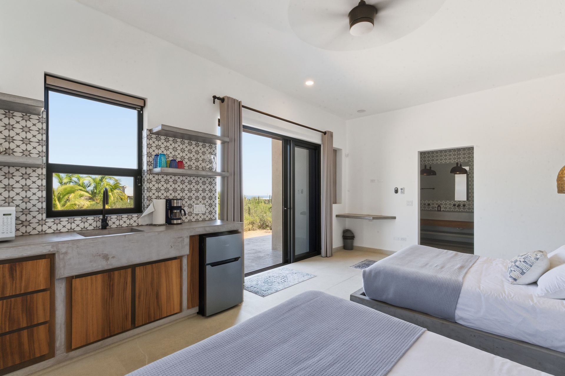 Modern hotel room with two beds, a small kitchenette with a sink, mini-fridge, and a sliding door leading to a balcony.