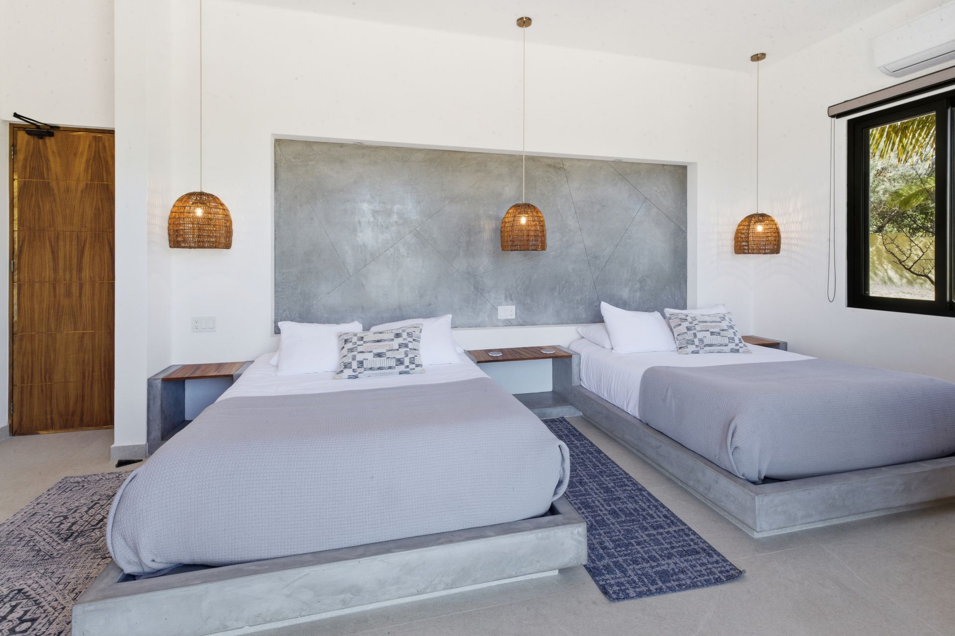A minimalist hotel room with two beds on grey concrete platforms, textured wall, and woven hanging lights against white.