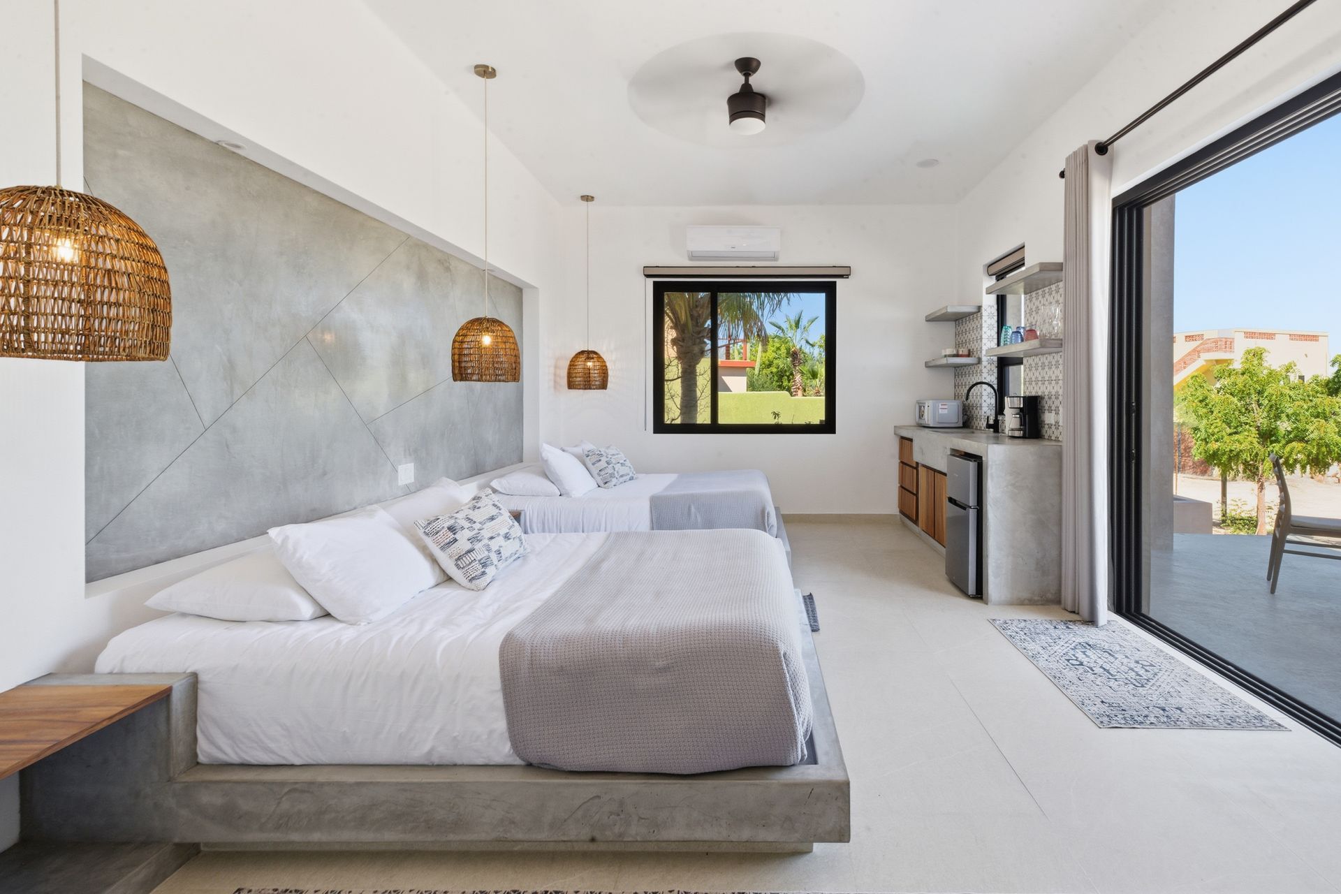 A modern hotel room with two white beds, concrete wall accents, woven pendant lights, and a sliding glass door to a patio.