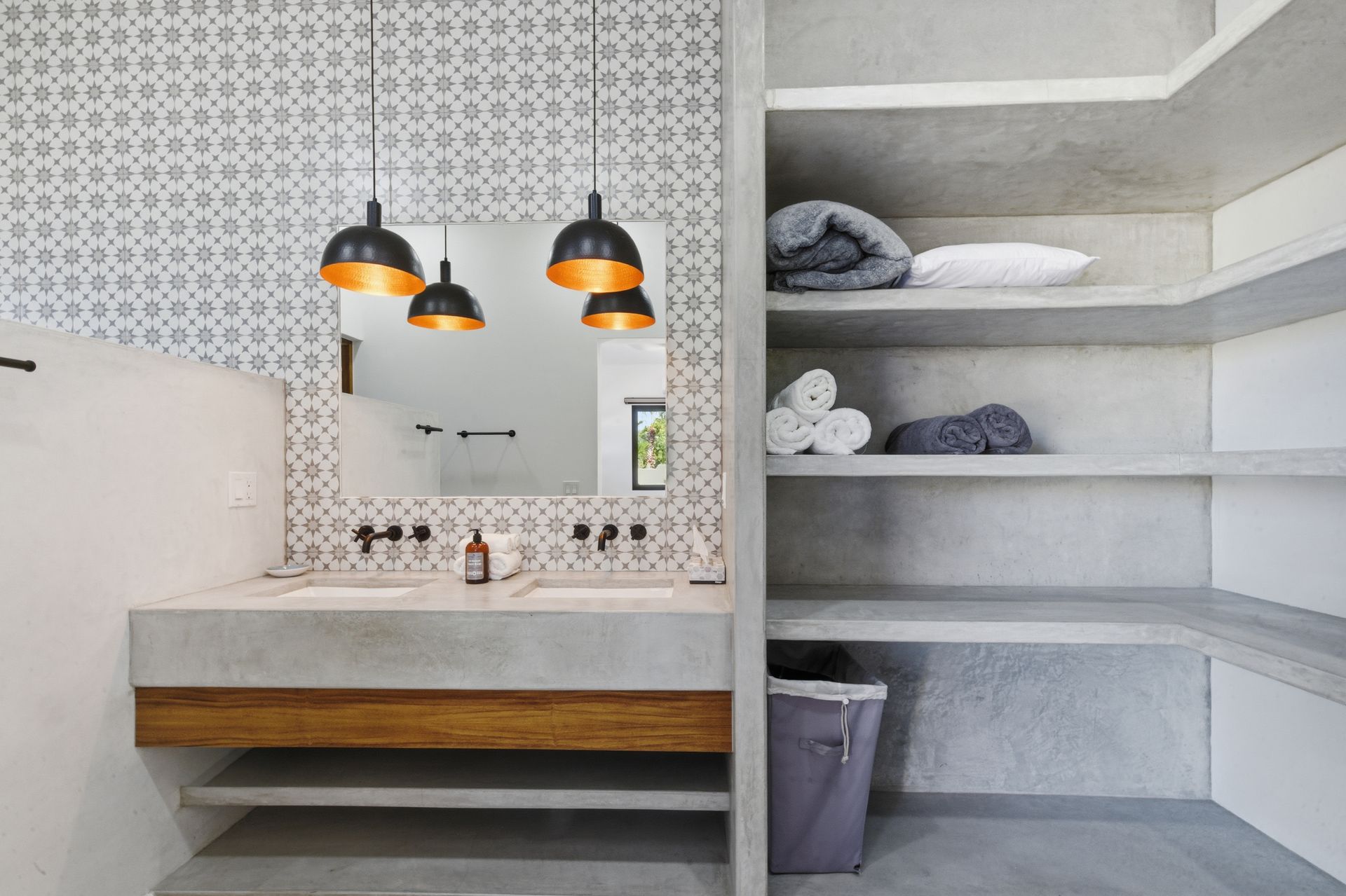 A modern bathroom featuring a concrete double vanity, patterned tile backsplash, pendant lights, and open shelving units.