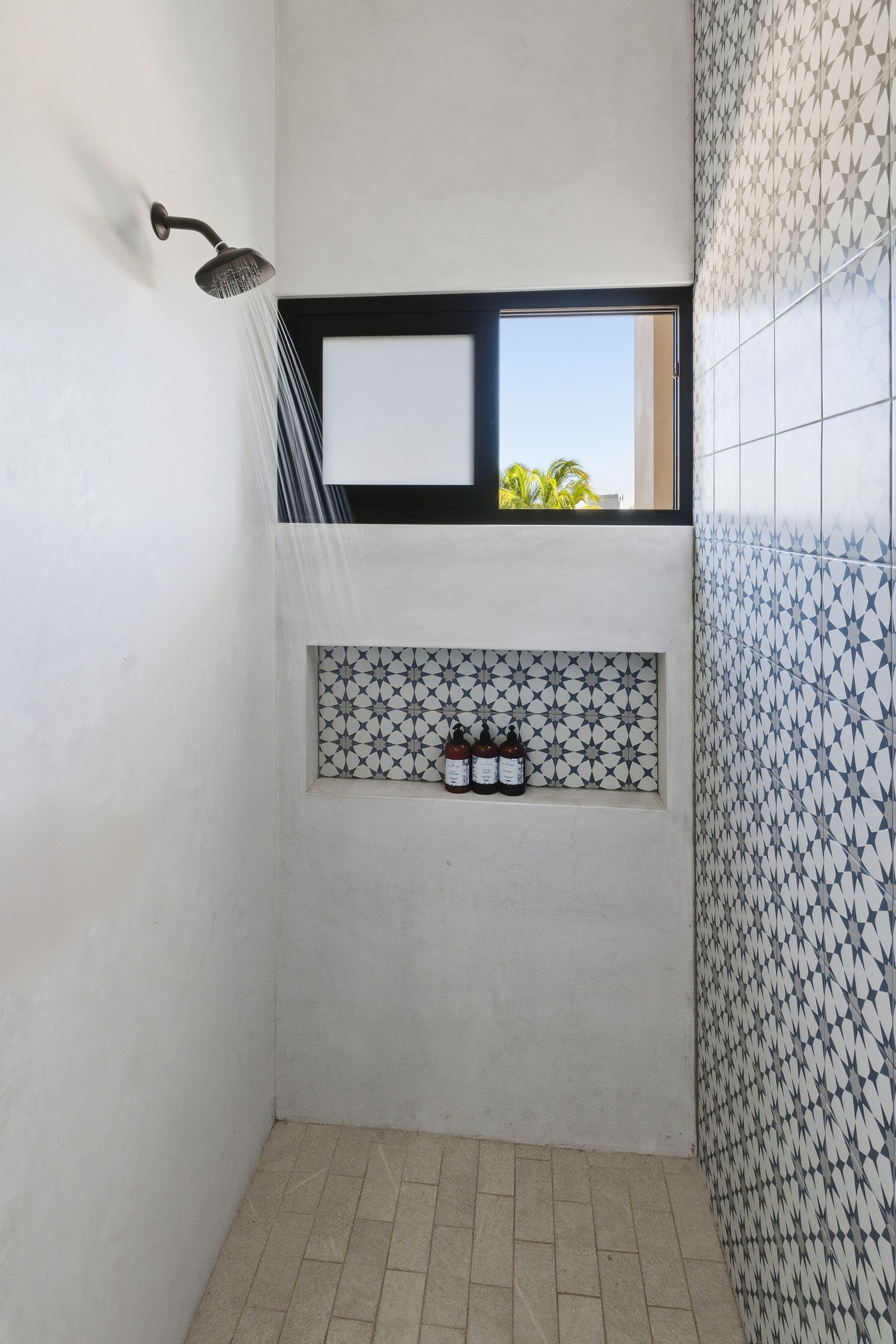 A modern shower stall with white walls, patterned tiles on one wall, a recessed shampoo niche, and a small square window.