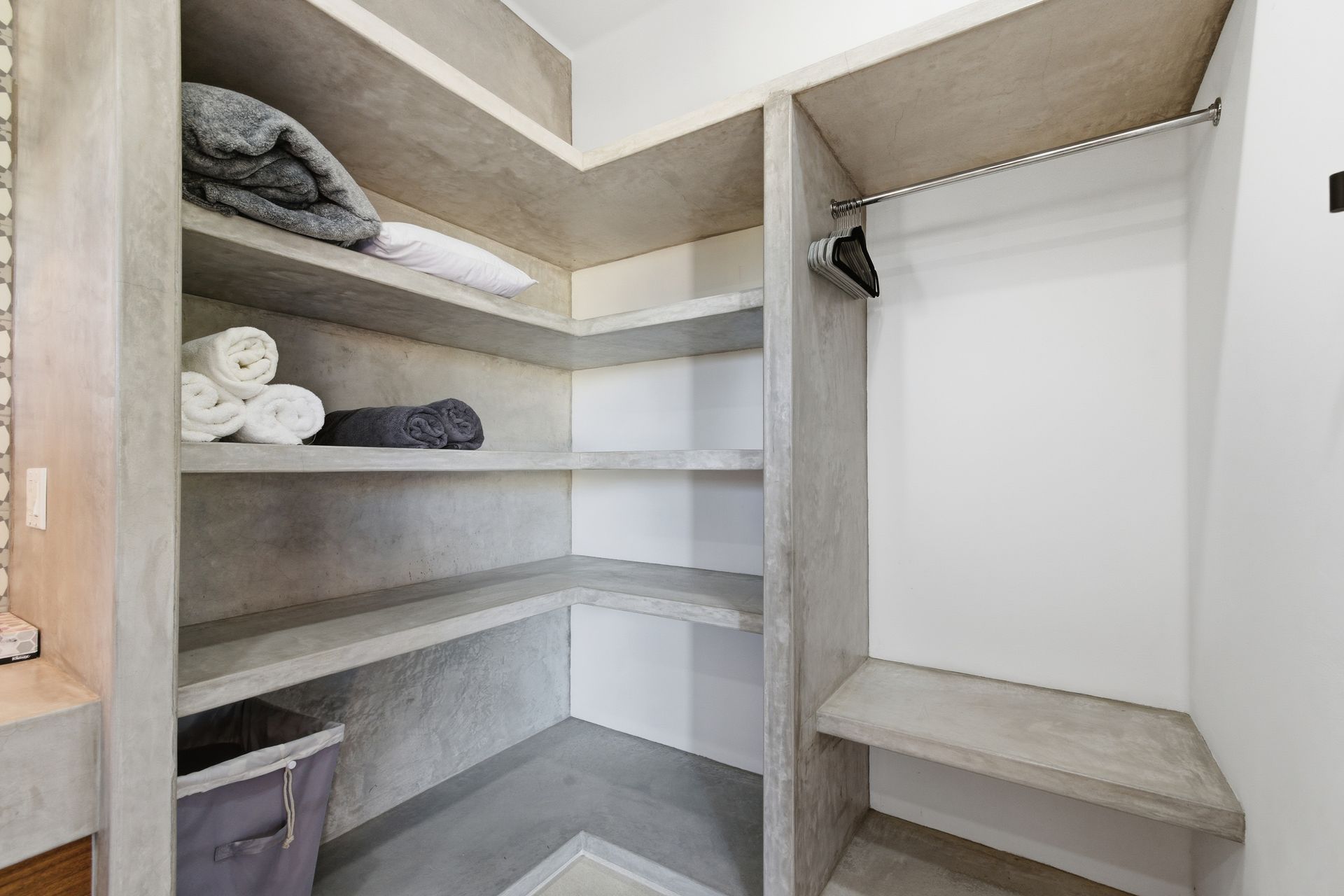 A modern walk-in closet with concrete shelves, white walls, stacked towels, a laundry hamper, and a clothes hanging rod.