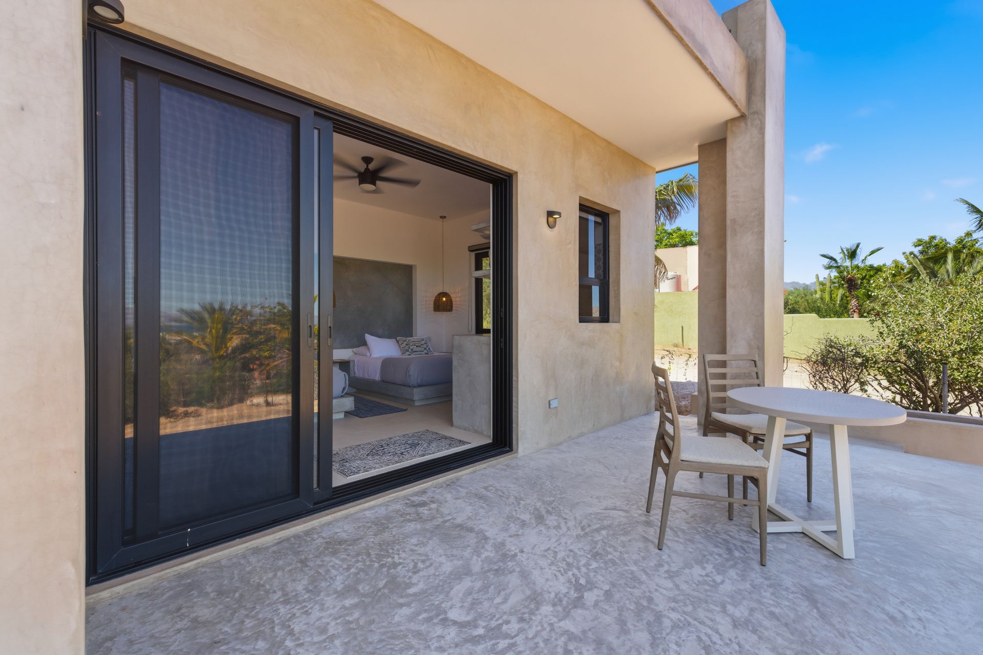 A patio with a small table and two chairs outside a room with sliding glass doors, overlooking a sunny landscape.