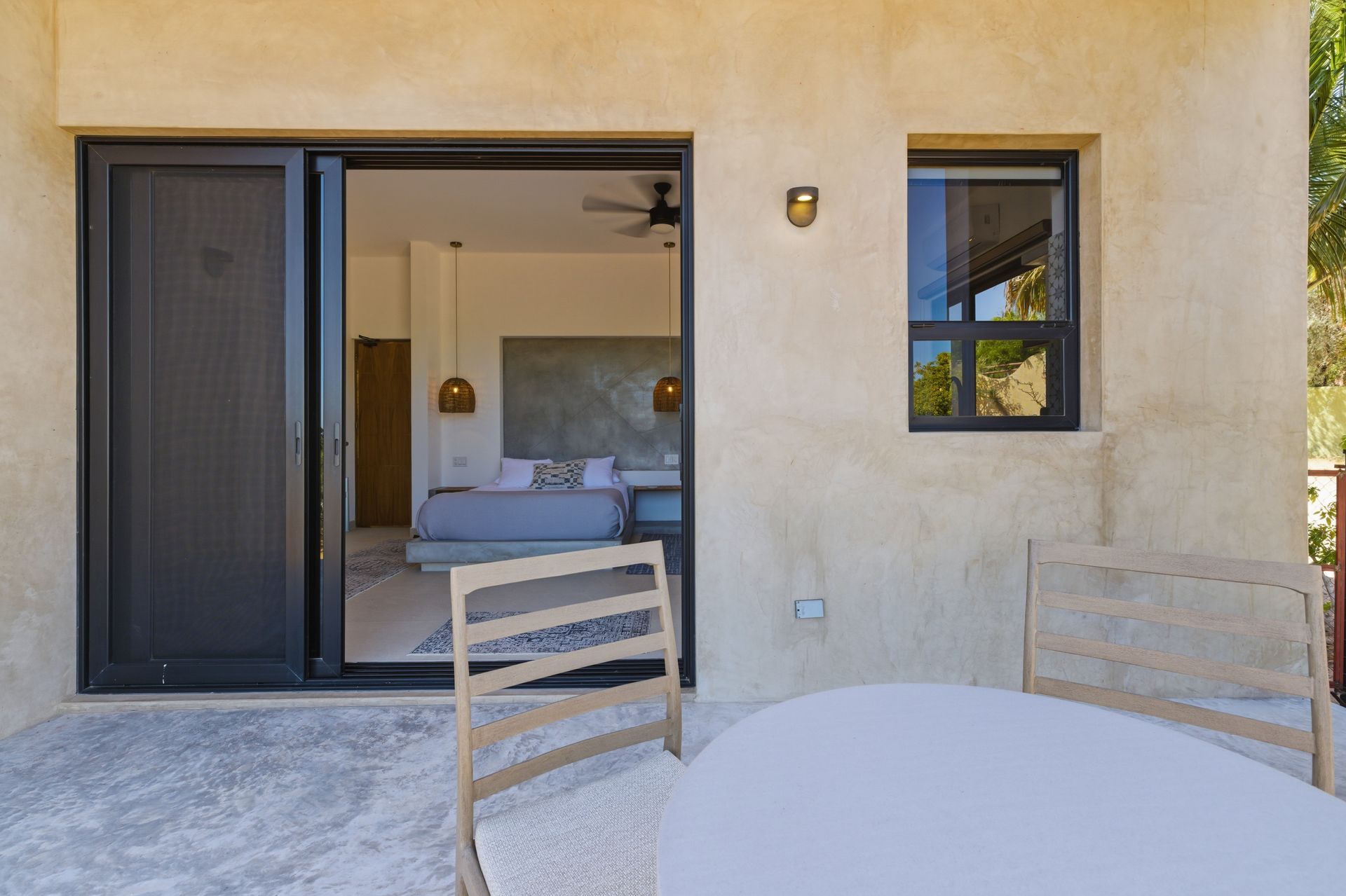 A beige stucco patio with a table and chairs, featuring a sliding glass door opening into a modern bedroom.