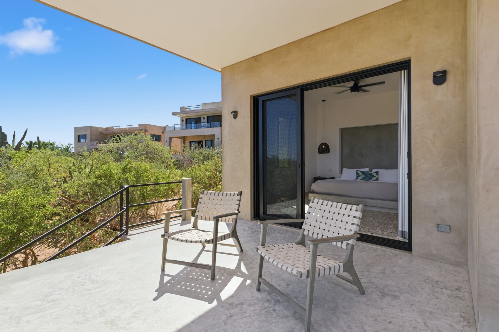 Two modern chairs on a stone patio overlooking green bushes under a bright blue sky, with a view into a bedroom.