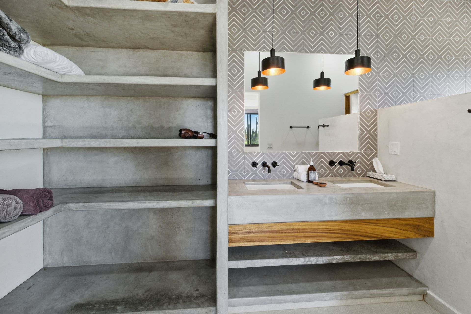 Modern bathroom with a dual concrete vanity, wood-paneled accent, gray geometric tile wall, and built-in concrete shelving.