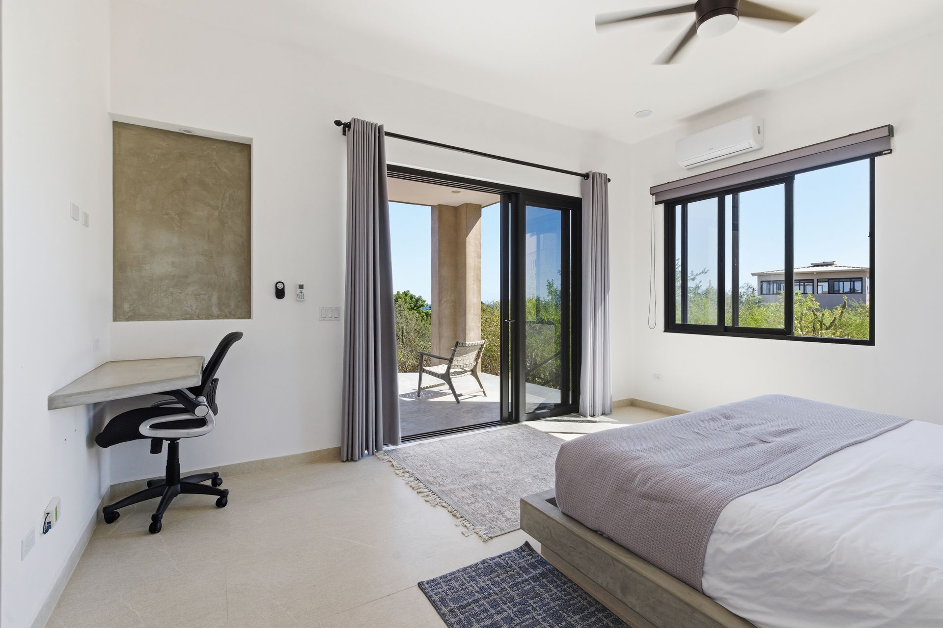 A bright bedroom with a bed, desk with a chair, and sliding glass doors leading to a patio with a chair.