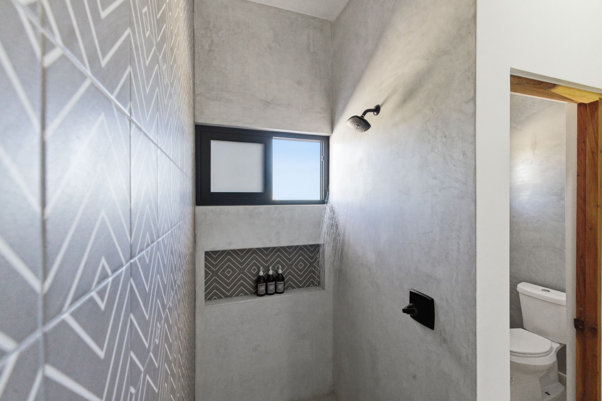 A shower with grey concrete walls, a patterned tile accent wall, a small window, and a built-in shampoo niche.